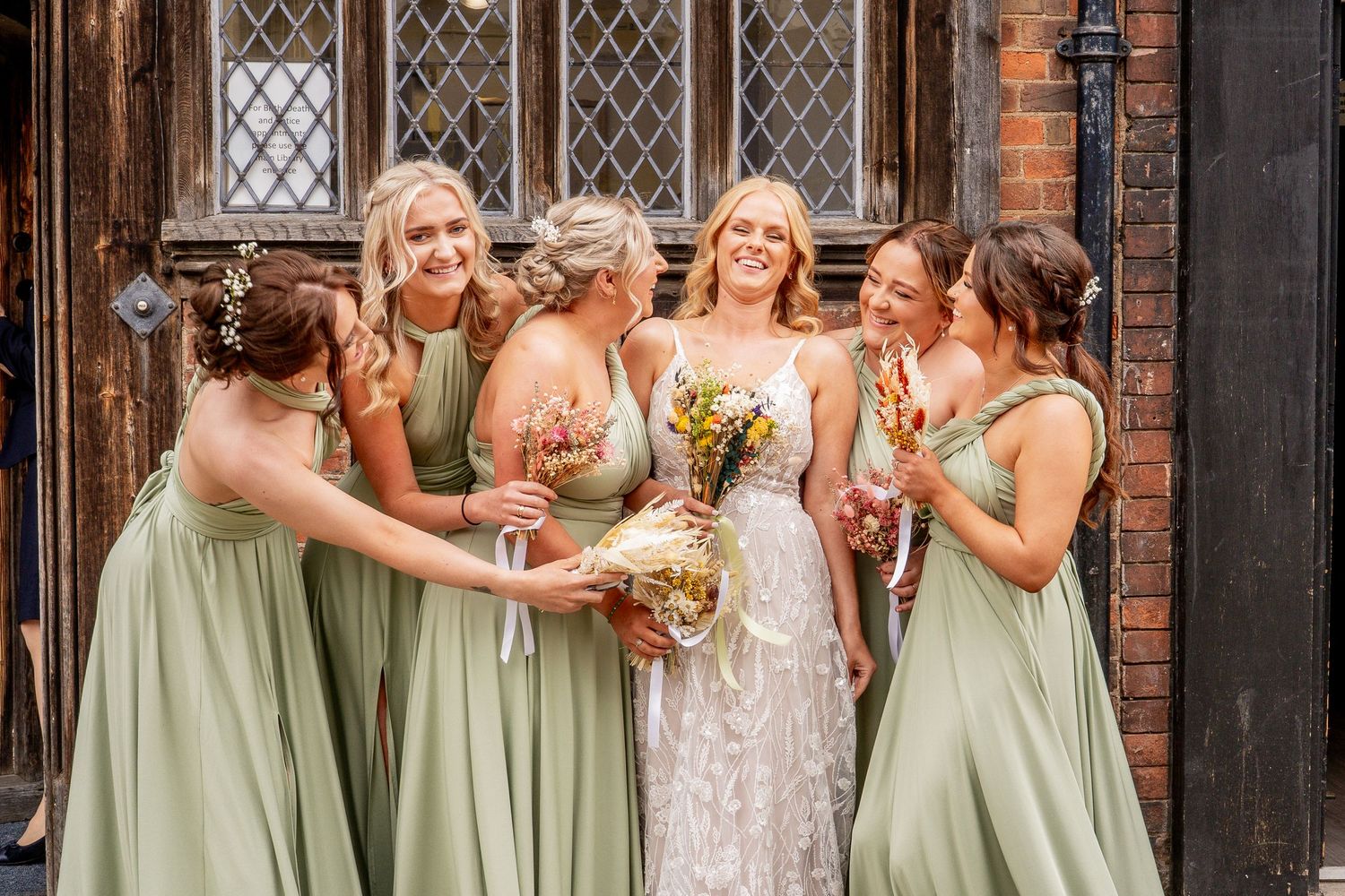 A wedding party in sage green bridesmaid dresses and white lace bridal gown share a joyful moment outside the henley rooms.