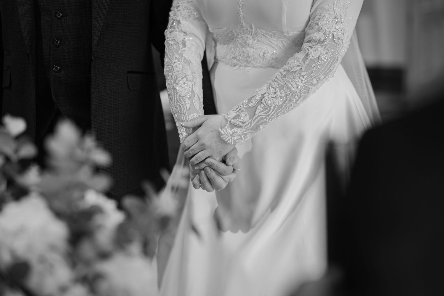 Elegant bride in a flowing white gown with lace sleeves clasps hands in front, captured in black and white photography.