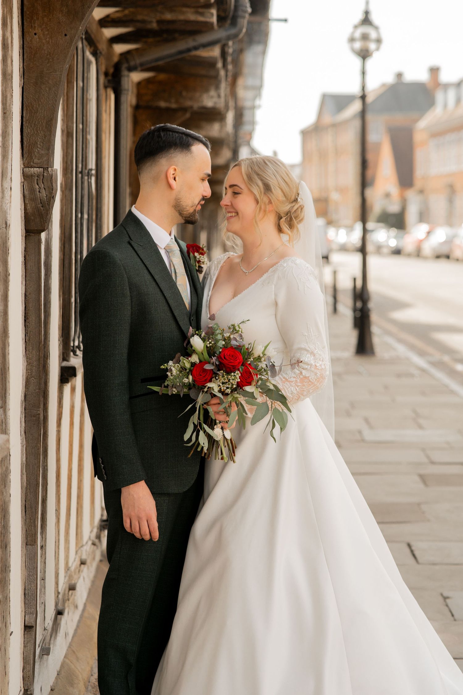 A bride and groom share an intimate moment on a historic street, with the bride holding a red and green bouquet.