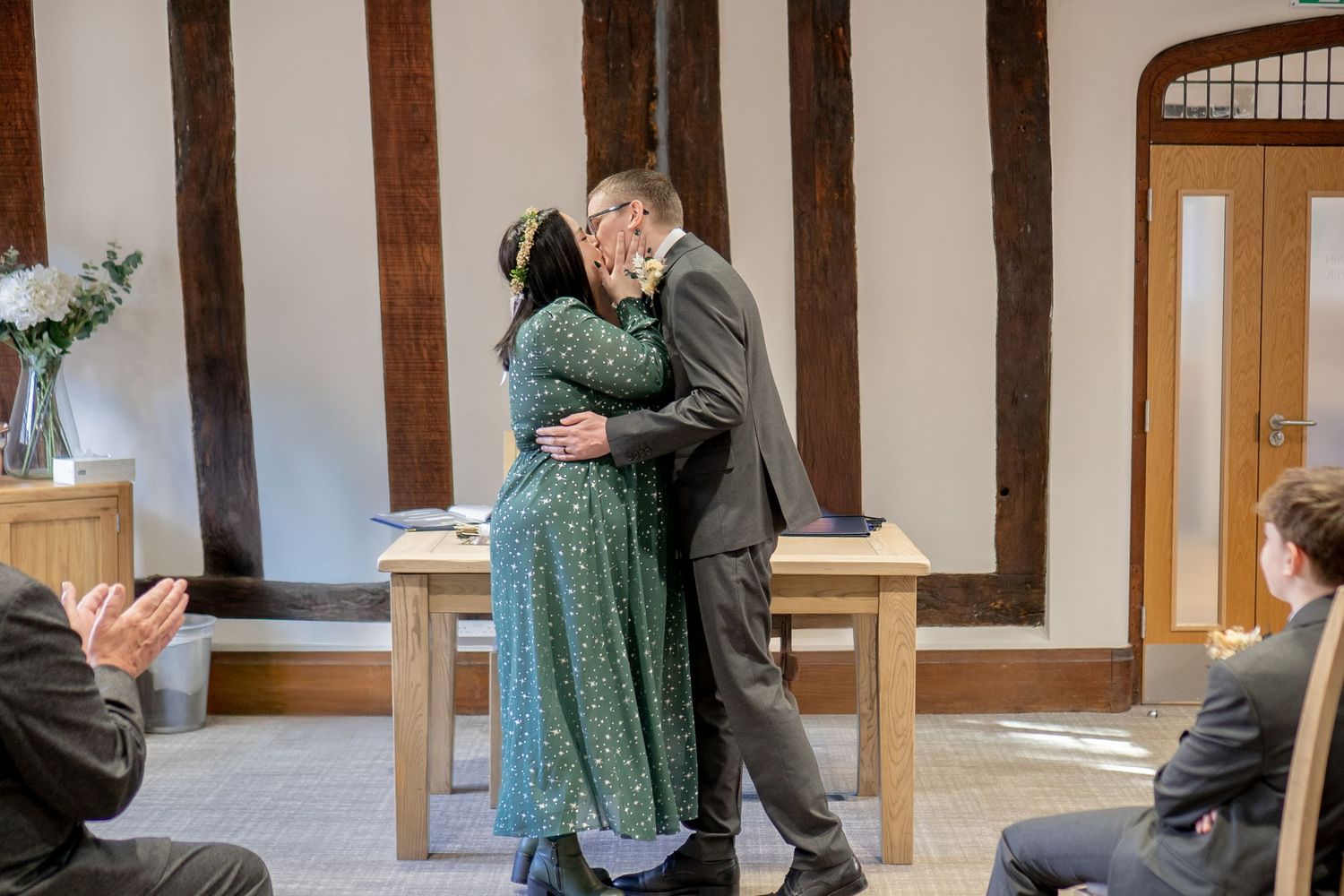 Two people embrace warmly during an intimate ceremony while onlookers applaud in a room with wooden beams and white walls.