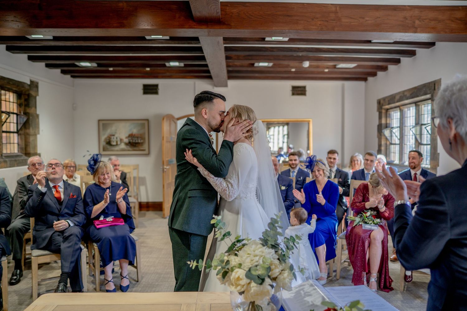 Wedding ceremony at an intimate venue with exposed wooden beams and guests watching the couple share their first kiss.