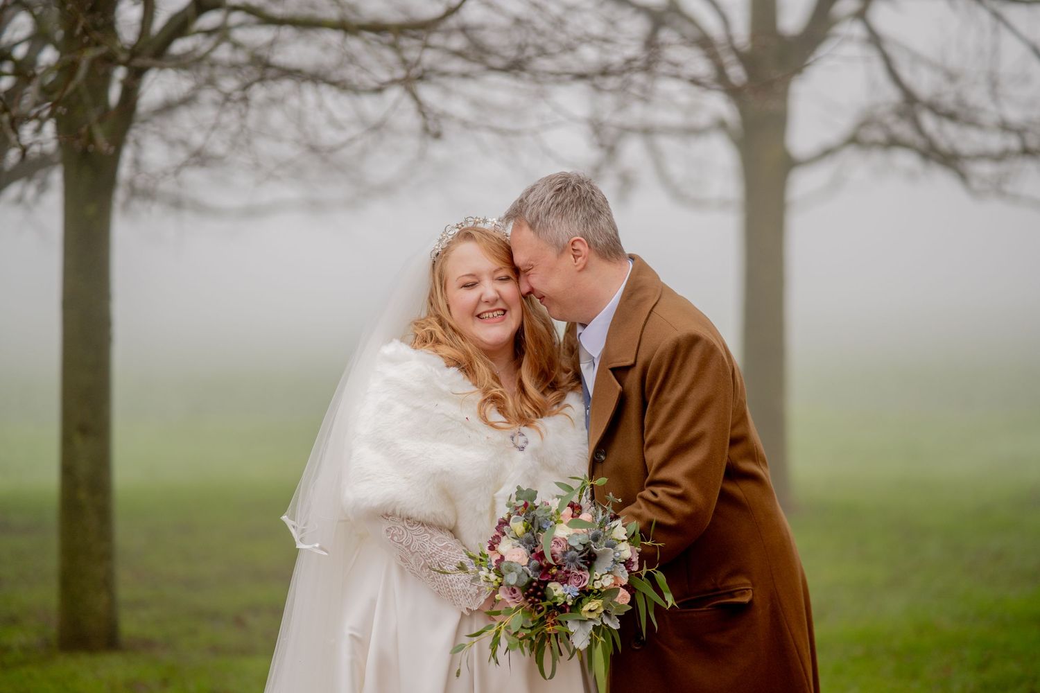 A romantic winter wedding couple shares a tender moment in the misty countryside while holding a rustic bouquet.