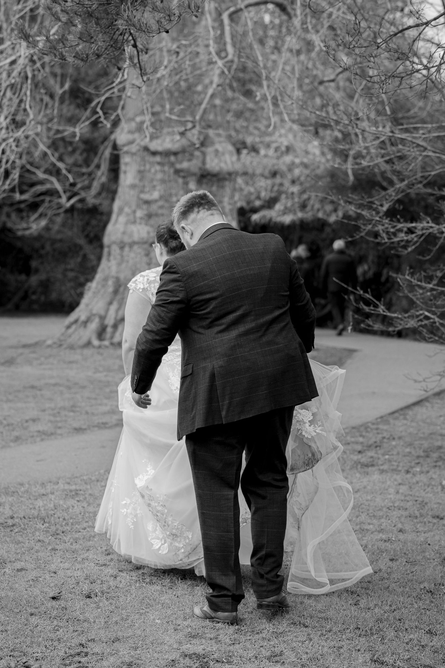 A tender black and white wedding moment captured outdoors with a bride in a flowing dress walking with her companion.