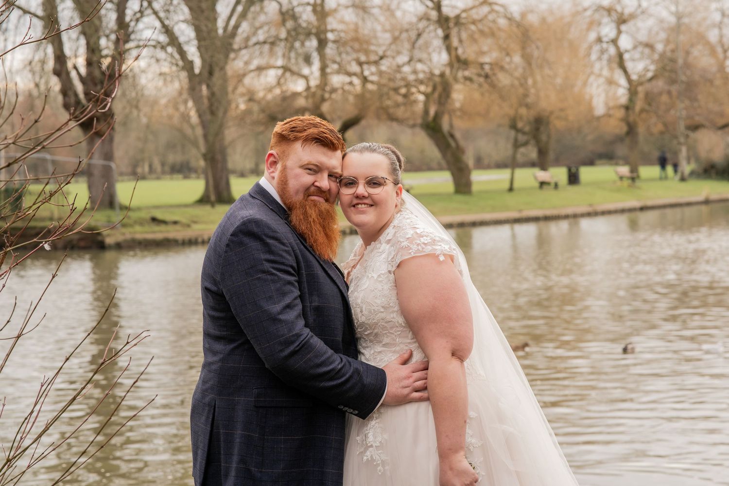 A couple embraces beside a serene pond in a park setting during autumn, with bare trees in the background.