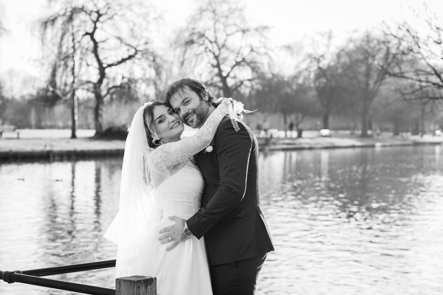 Newlywed couple embraces in romantic pose by serene riverside on their wedding day in black and white photography.