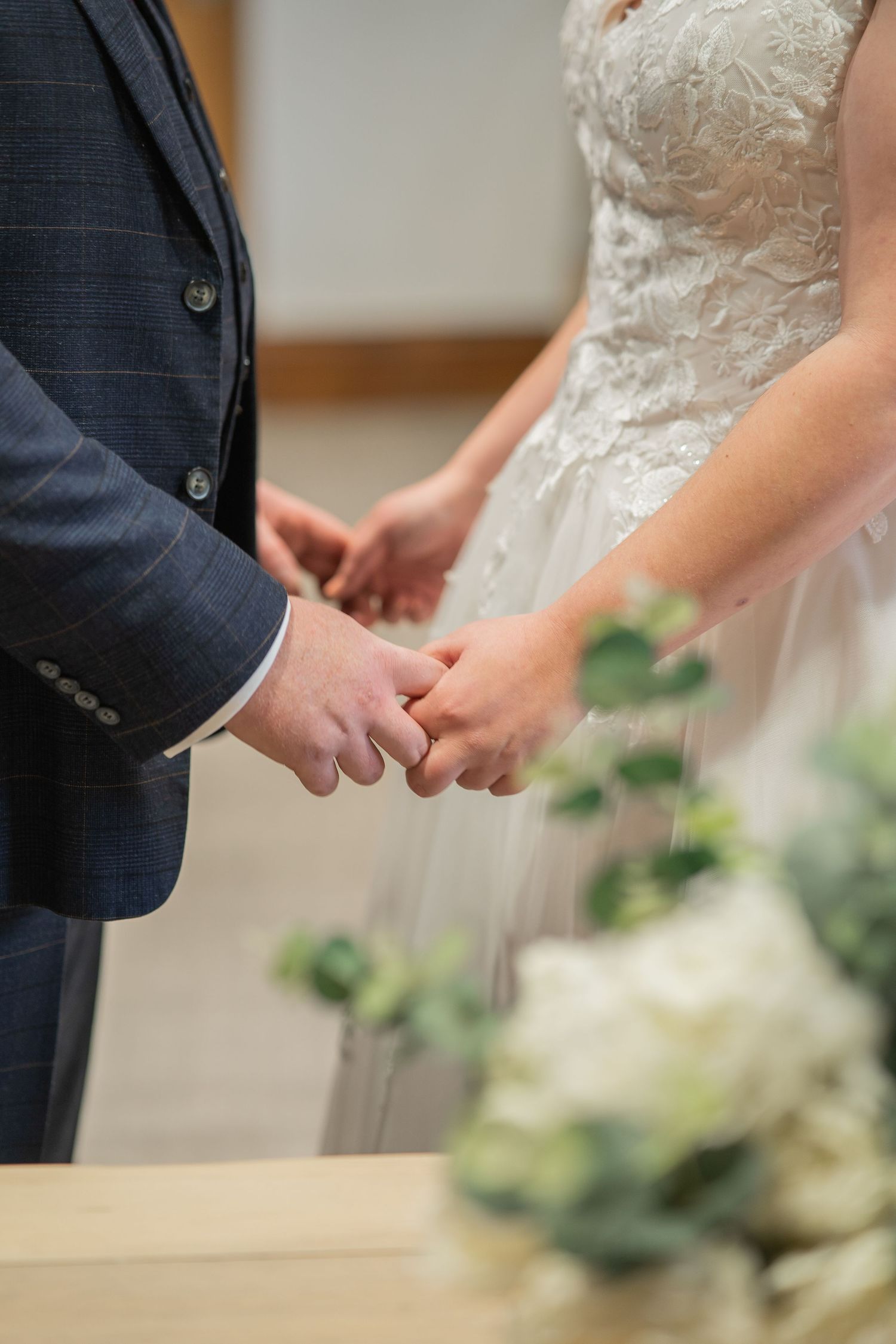 Close-up of couple holding hands during wedding ceremony with white floral decor and navy suit visible.