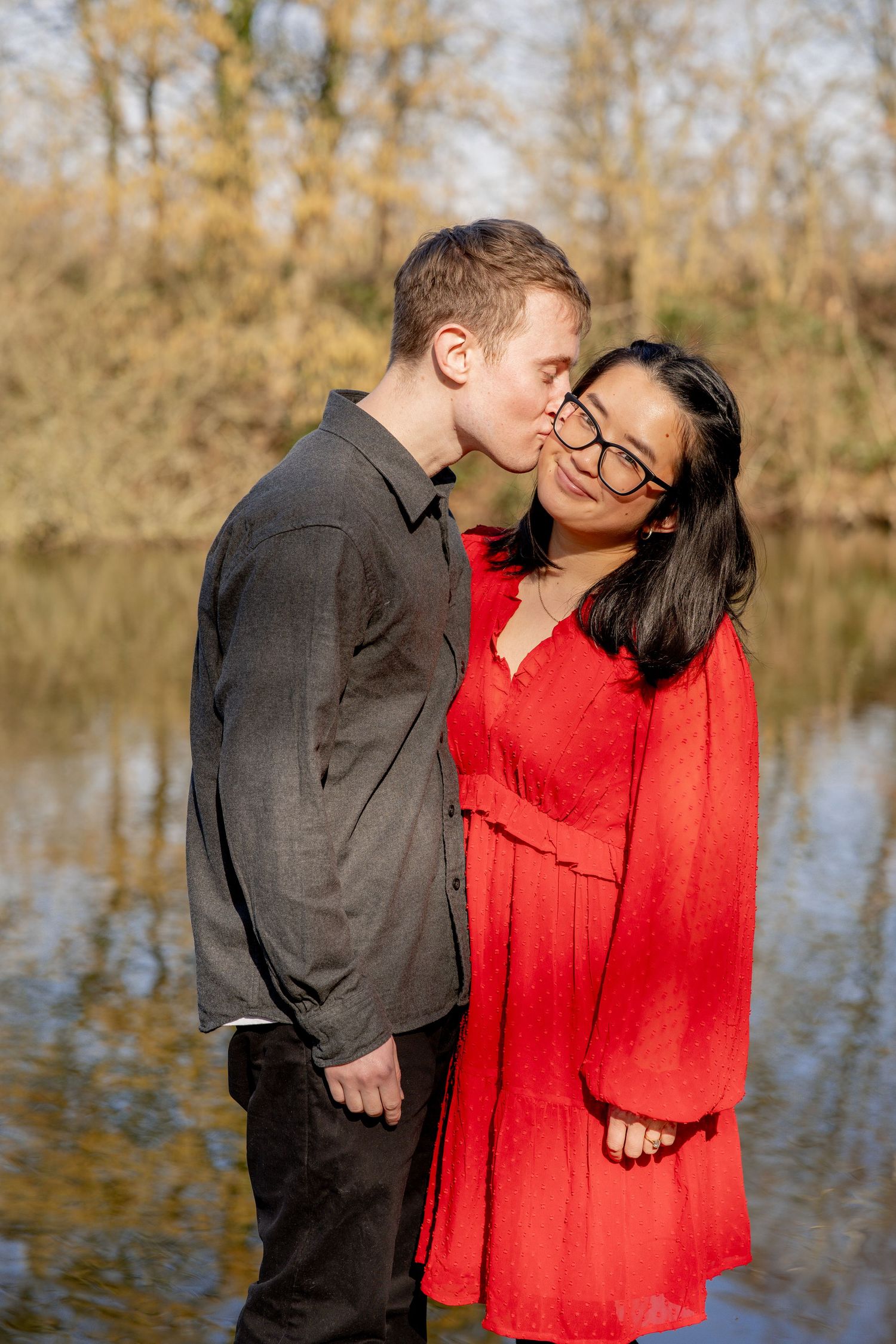A couple shares a tender moment by a lake on an autumn day, with one wearing a red dress and the other in dark attire.