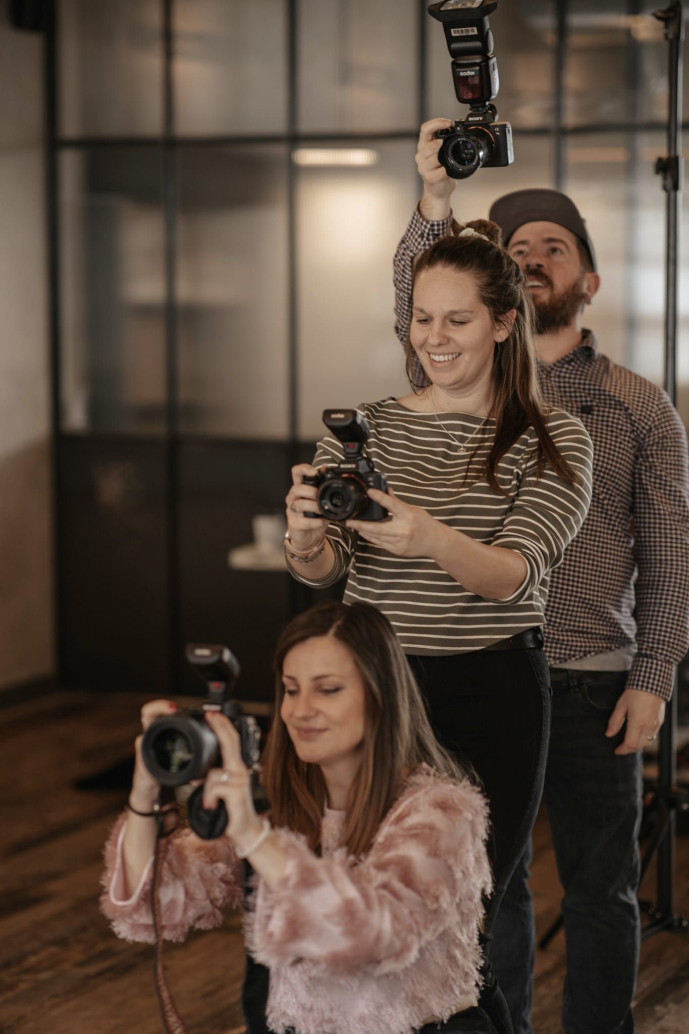 Photographers with professional cameras work together in indoor studio space with natural lighting and wooden floors.
