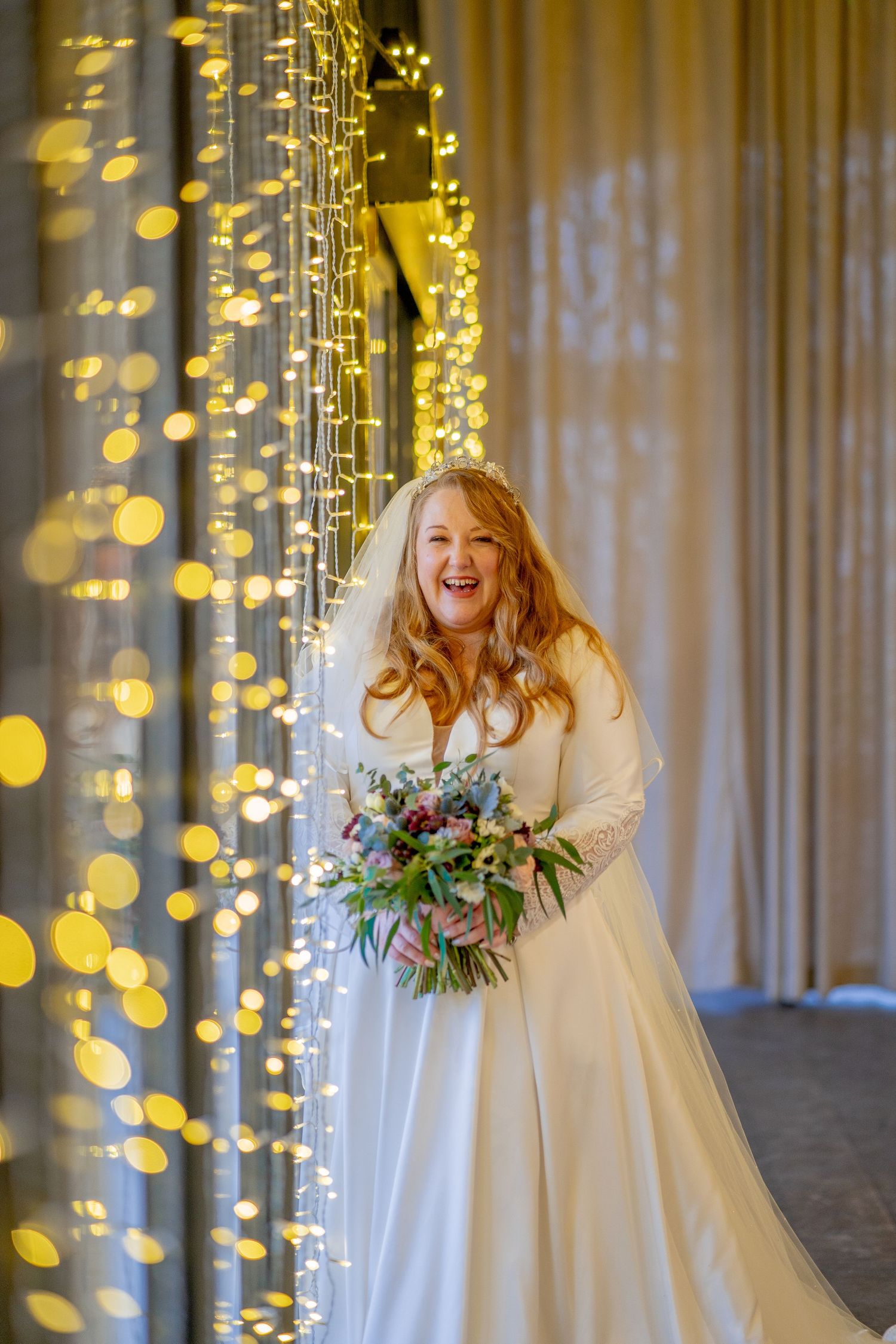 A bride in a white wedding gown holds a bouquet while standing against a backdrop of twinkling golden fairy lights.