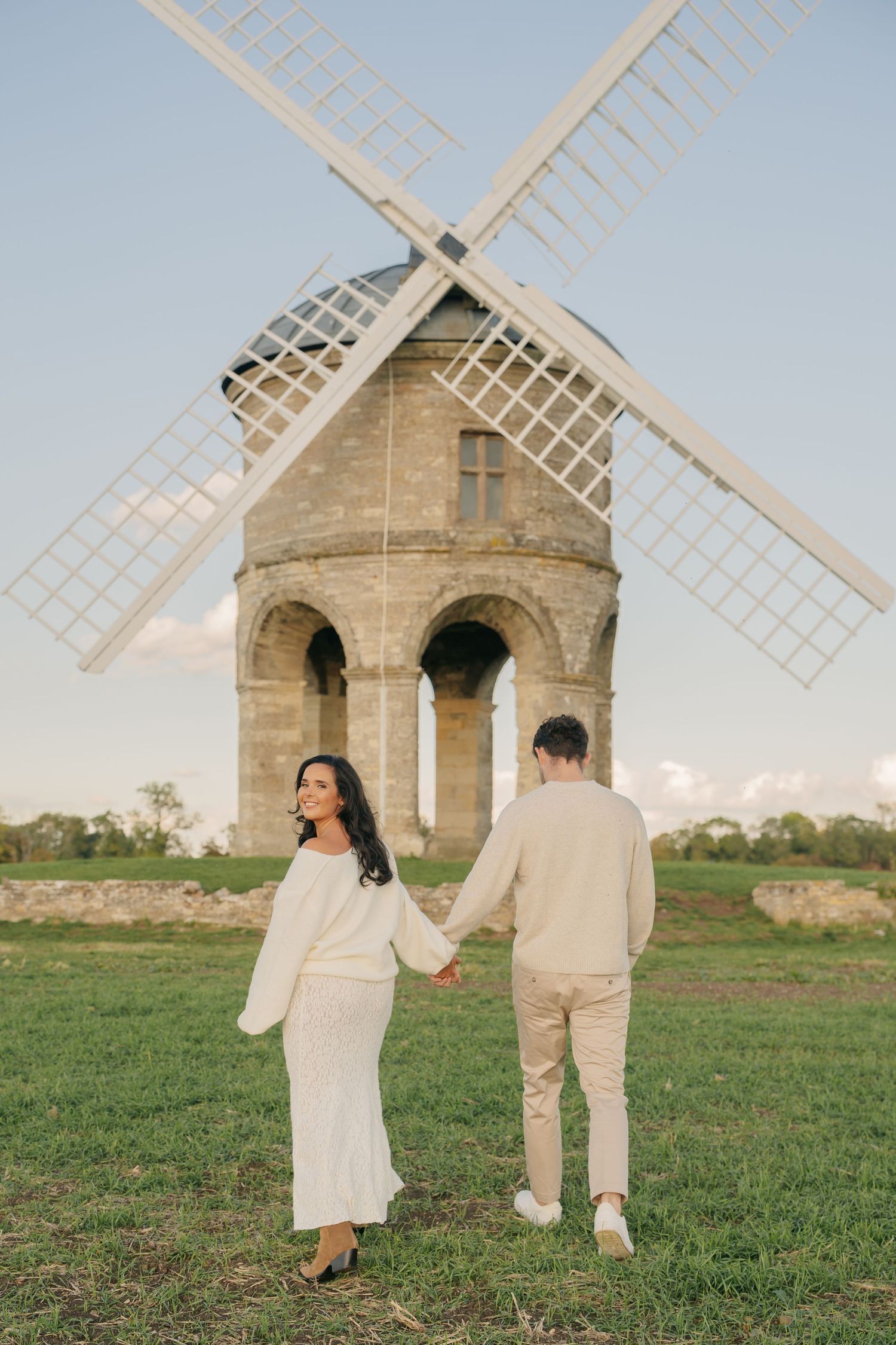 A couple in white outfits walks hand in hand near a historic stone windmill in a grassy field at sunset.