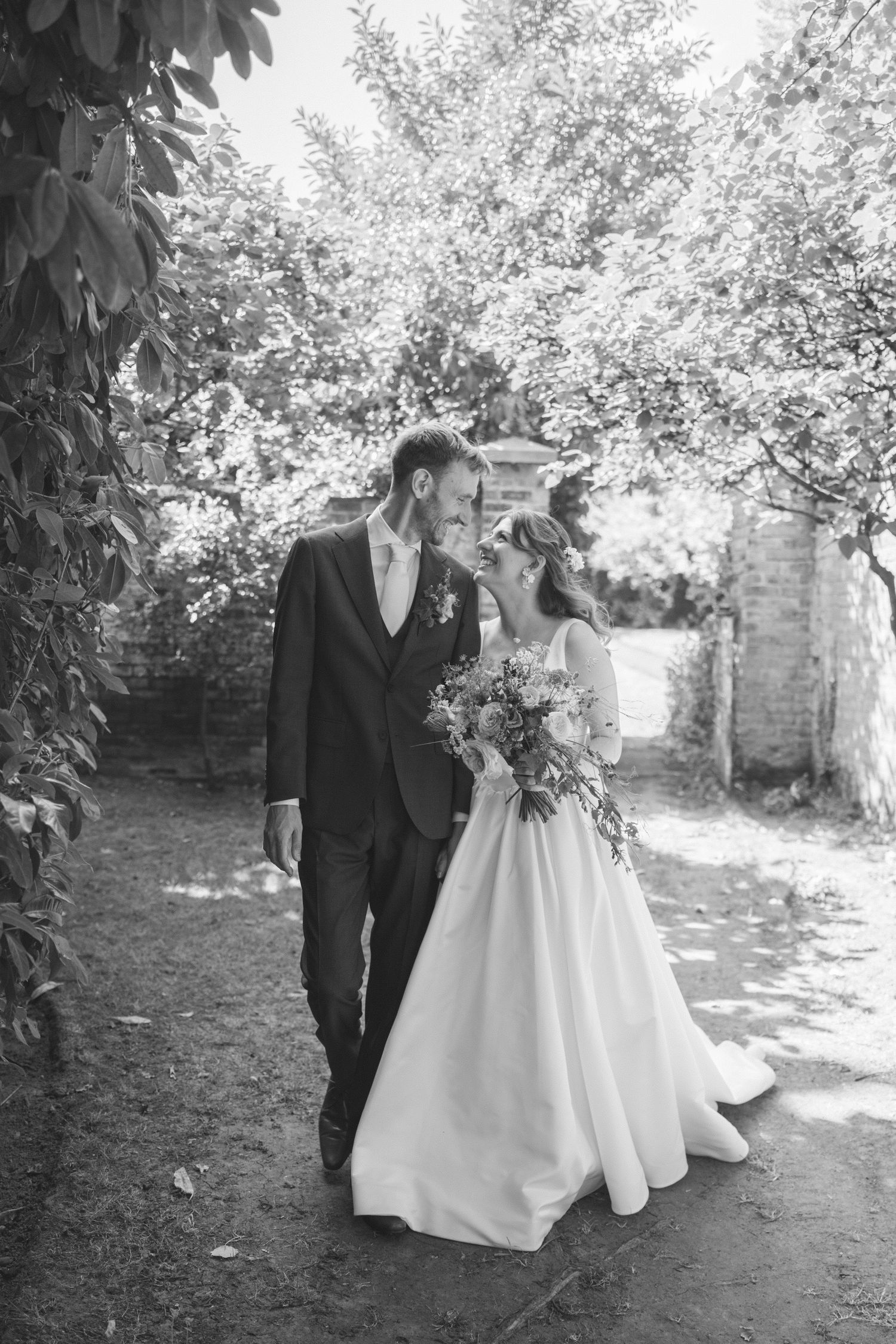 Black and white photo of a bride and groom walking together in a garden, smiling at each other, bride holding a bouquet.