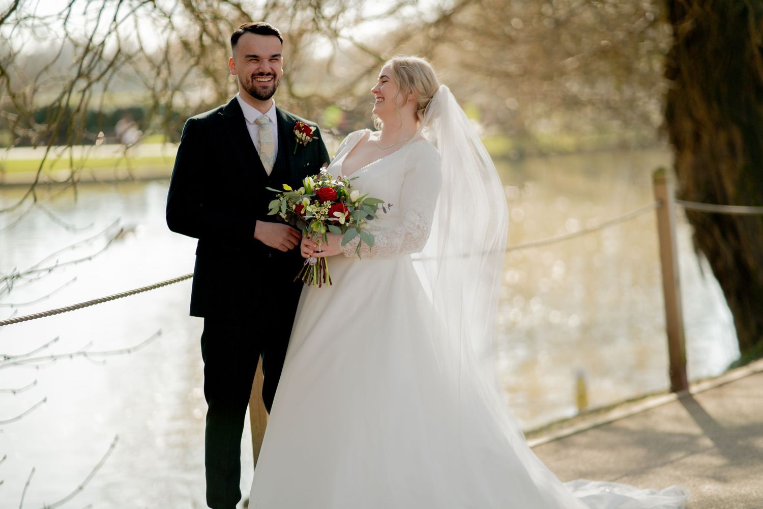 A newlywed couple in traditional wedding attire stands on a path near a lake on their wedding day.