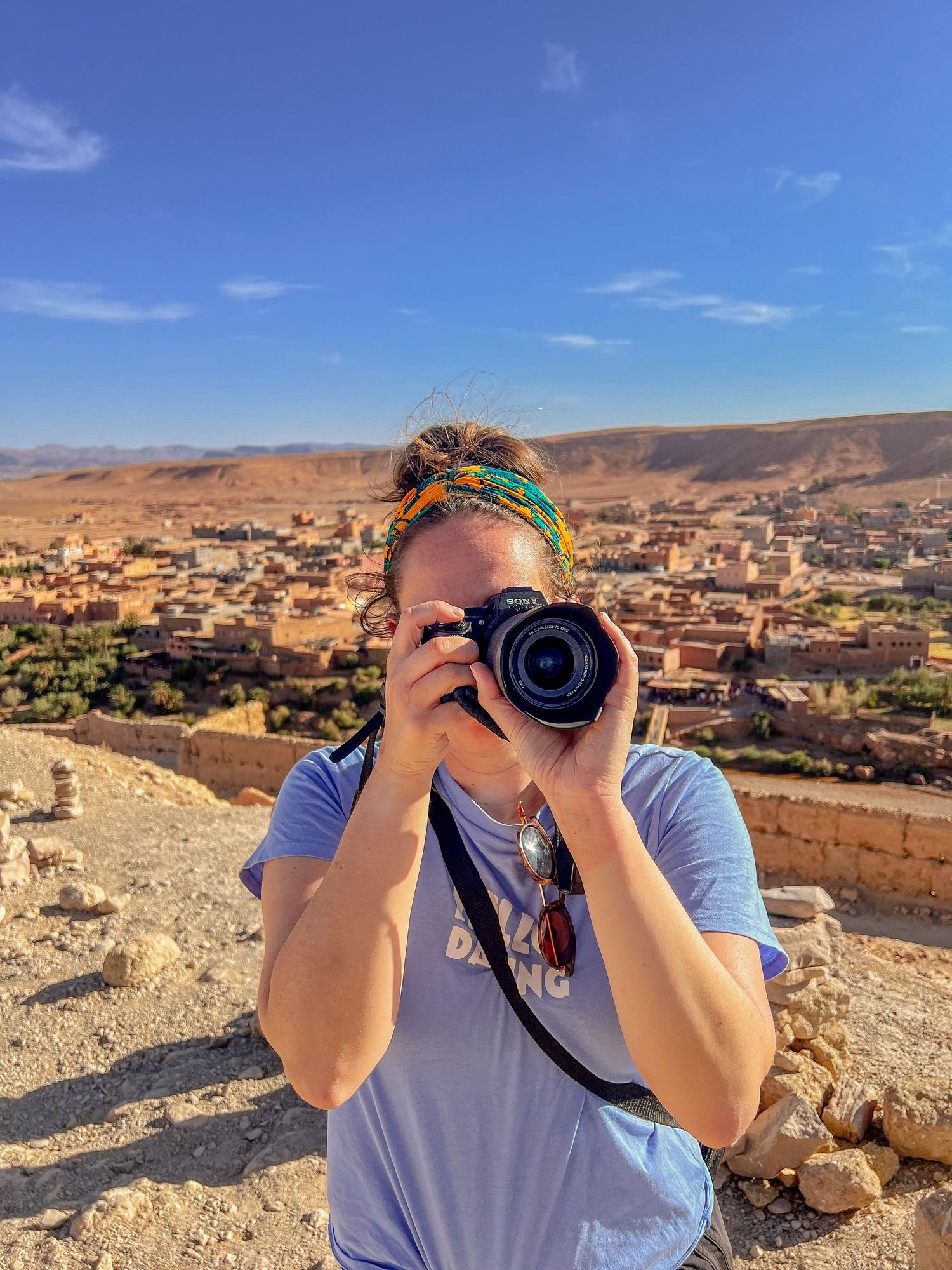 A photographer with a colorful headscarf captures images from a hilltop overlooking a desert town in Morocco.