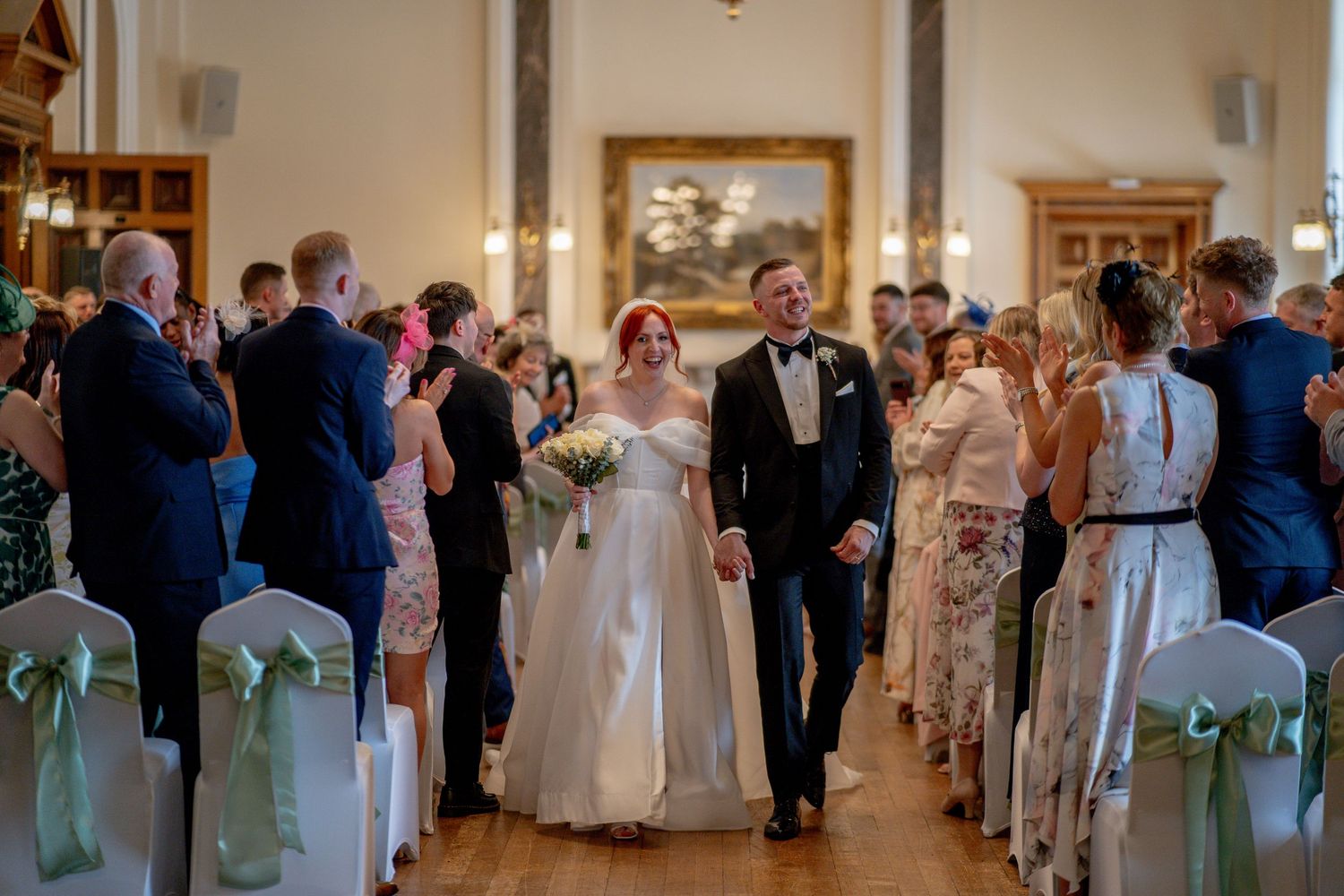 A wedding ceremony processional in an elegant indoor venue with white chair decorations and smiling guests looking on.