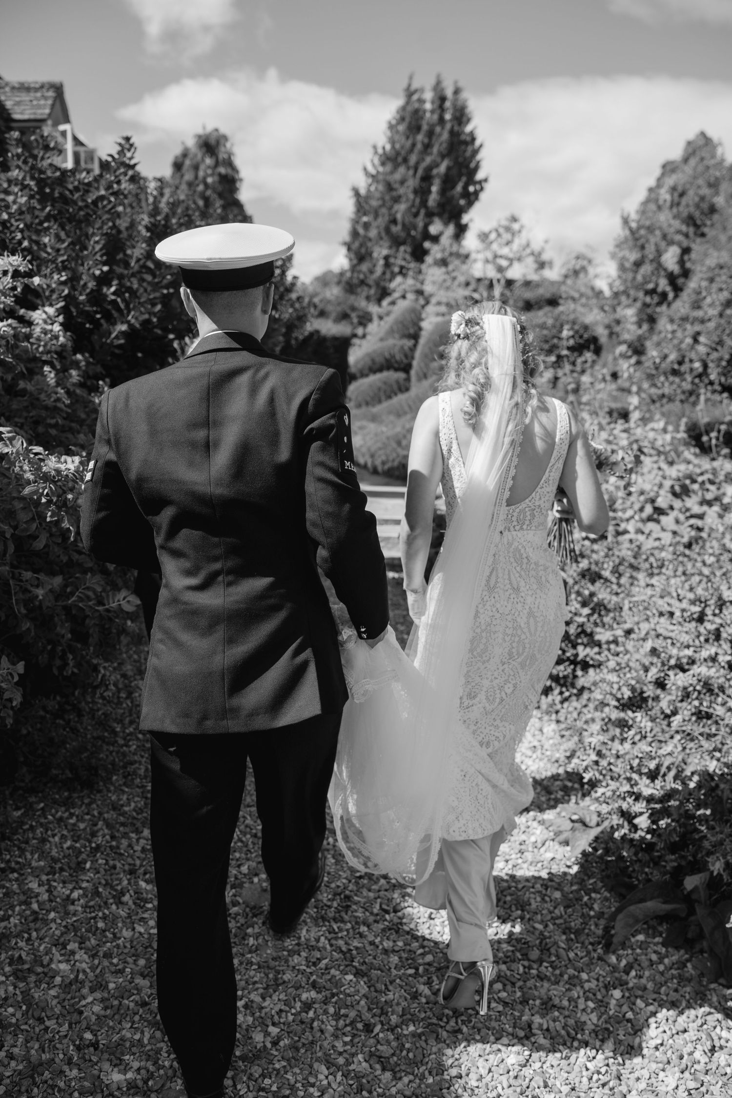 Black and white photo of a couple in wedding attire walking through a garden, viewed from behind.