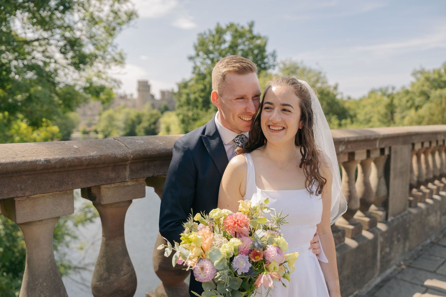 Bride and groom smiling together on a stone bridge, bride holding a colorful bouquet, lush green trees in the background.