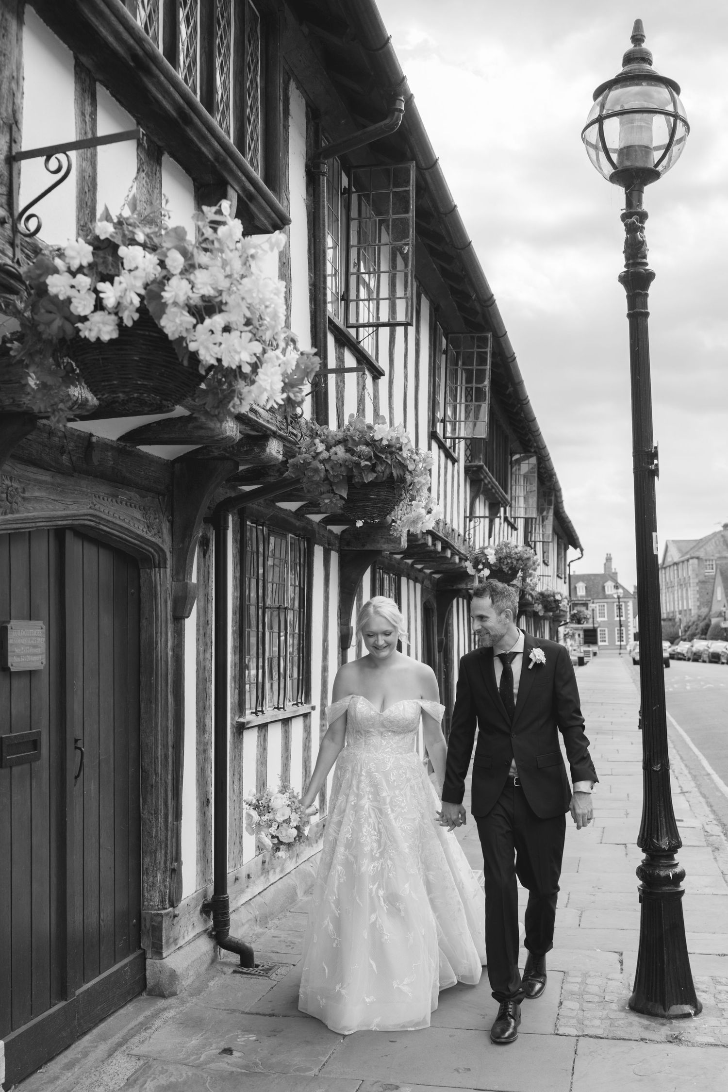 Black and white photo of a bride and groom walking hand in hand past a historic Tudor building adorned with floral arrangements.