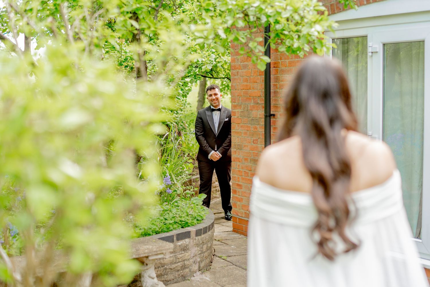 First look wedding moment captured in a garden setting with brick walls and greenery.