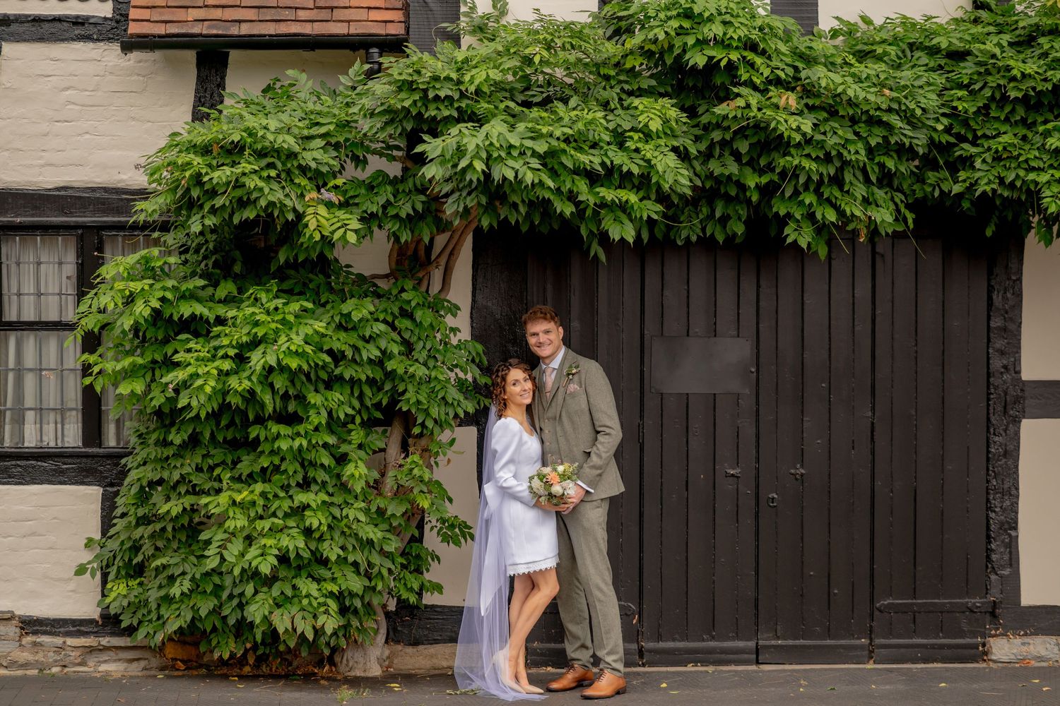 A couple poses together against a dark wall with lush green foliage growing overhead in an intimate moment.