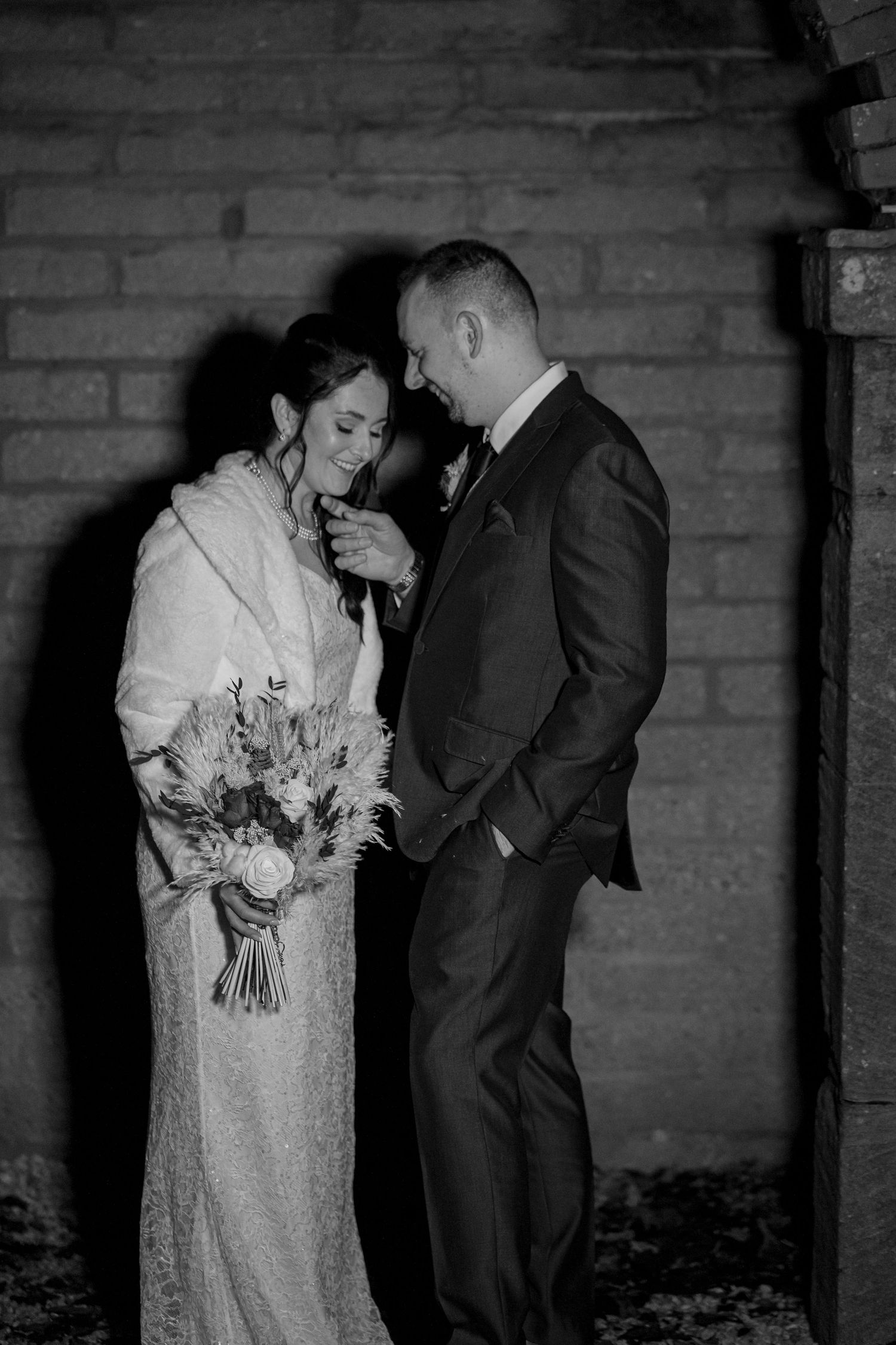 Black and white wedding photo showing a tender moment between newlyweds against a brick wall background.