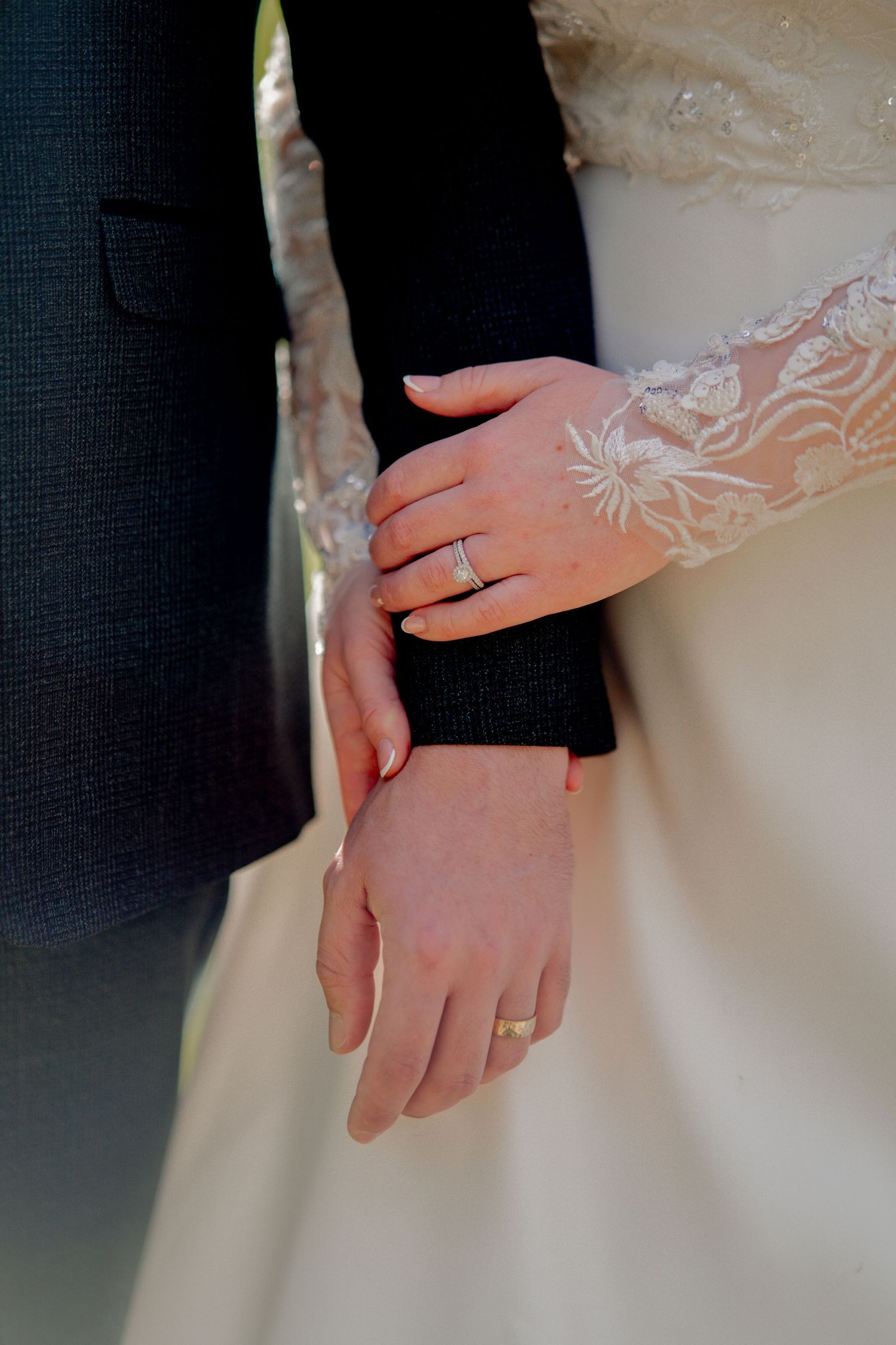 Close-up of bride and groom's hands touching, showing wedding rings and delicate lace sleeve details.