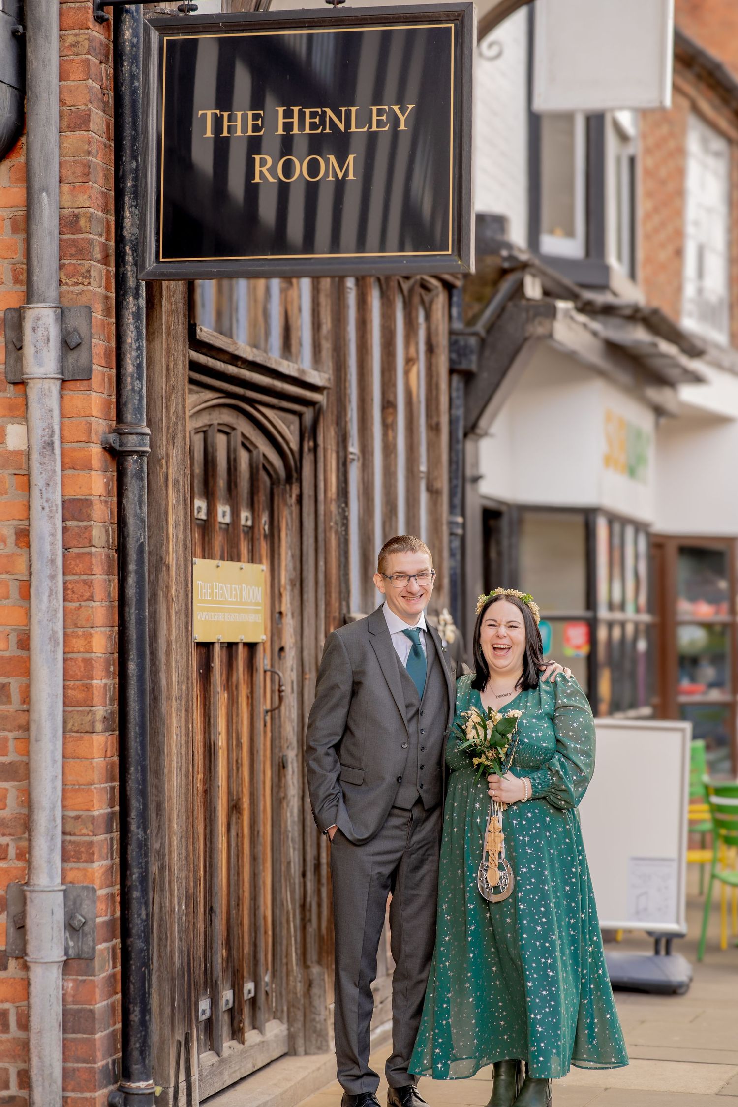 A couple stands outside The Henley Room entrance, with the woman wearing an emerald green dress and holding flowers.