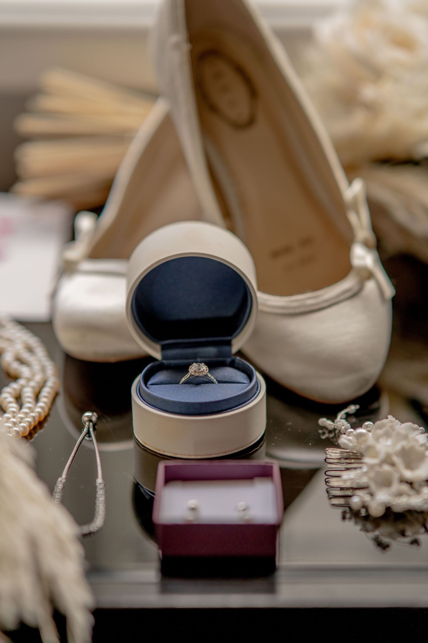 Wedding accessories arranged with white ballet flats, diamond engagement ring in navy box, and delicate lace details.