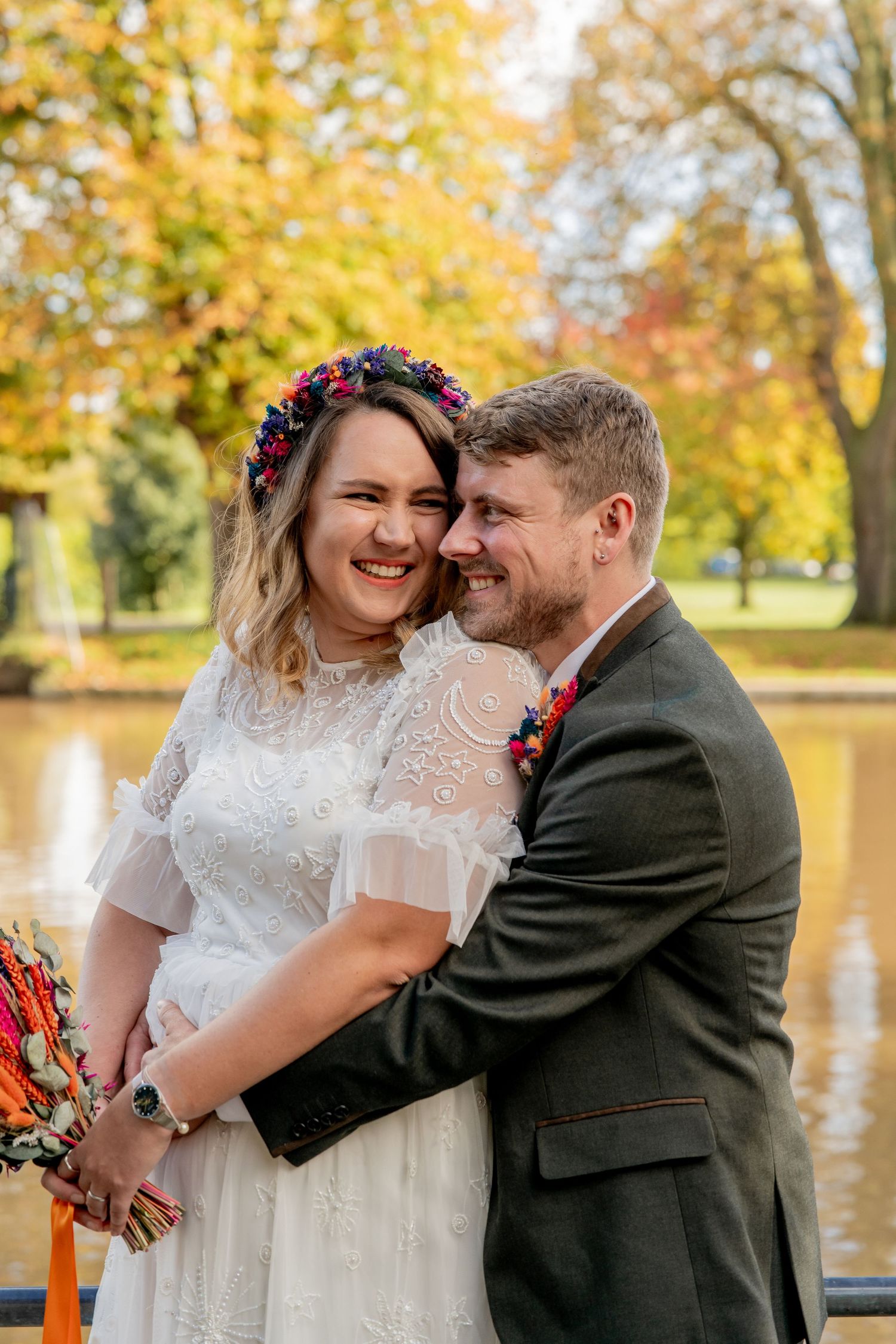 A joyful autumn wedding portrait with a bride in lace dress and flower crown embracing her partner by the river Avon.