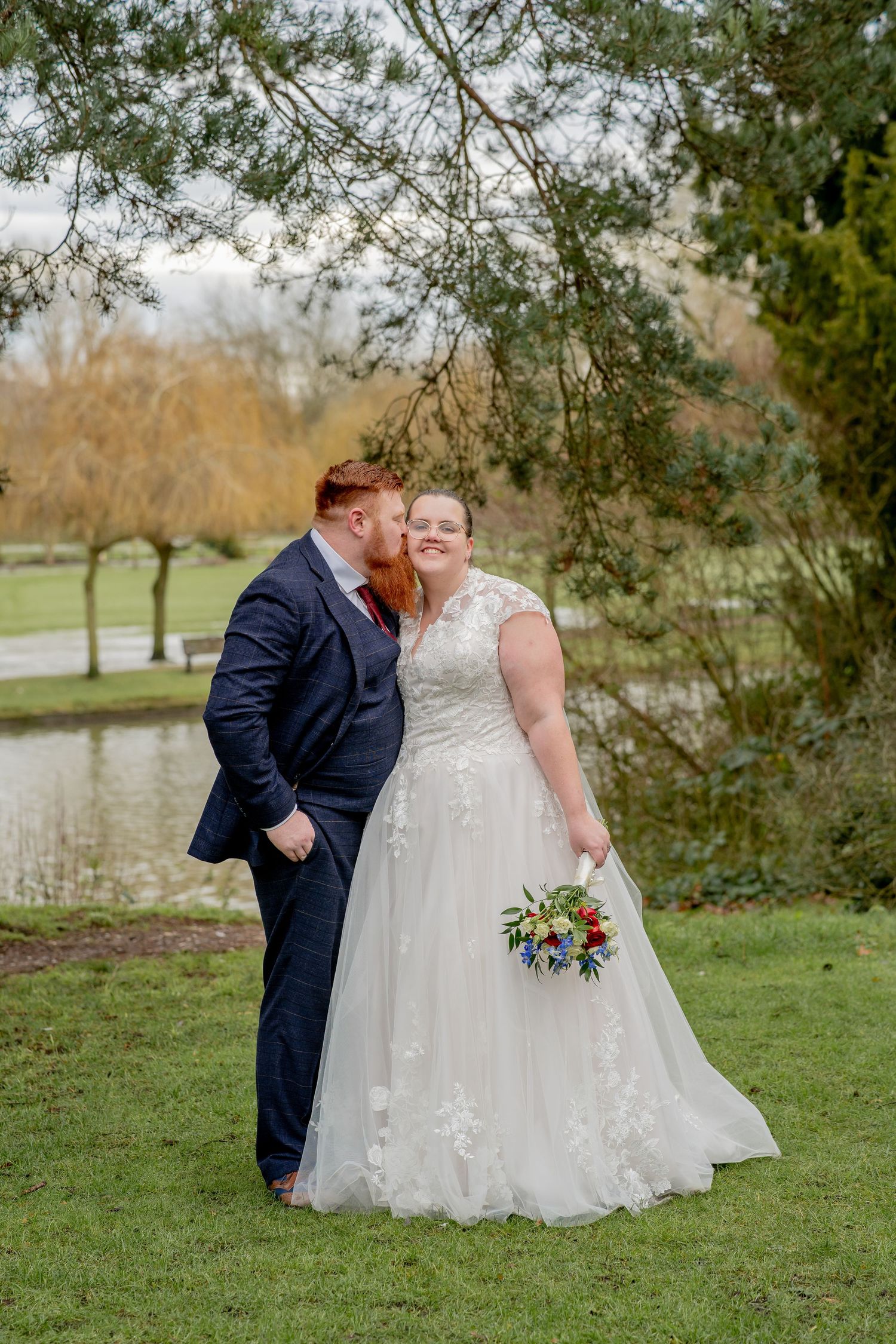 A romantic wedding moment captured outdoors near a pond with trees, featuring a navy suit and white lace gown with flowers.
