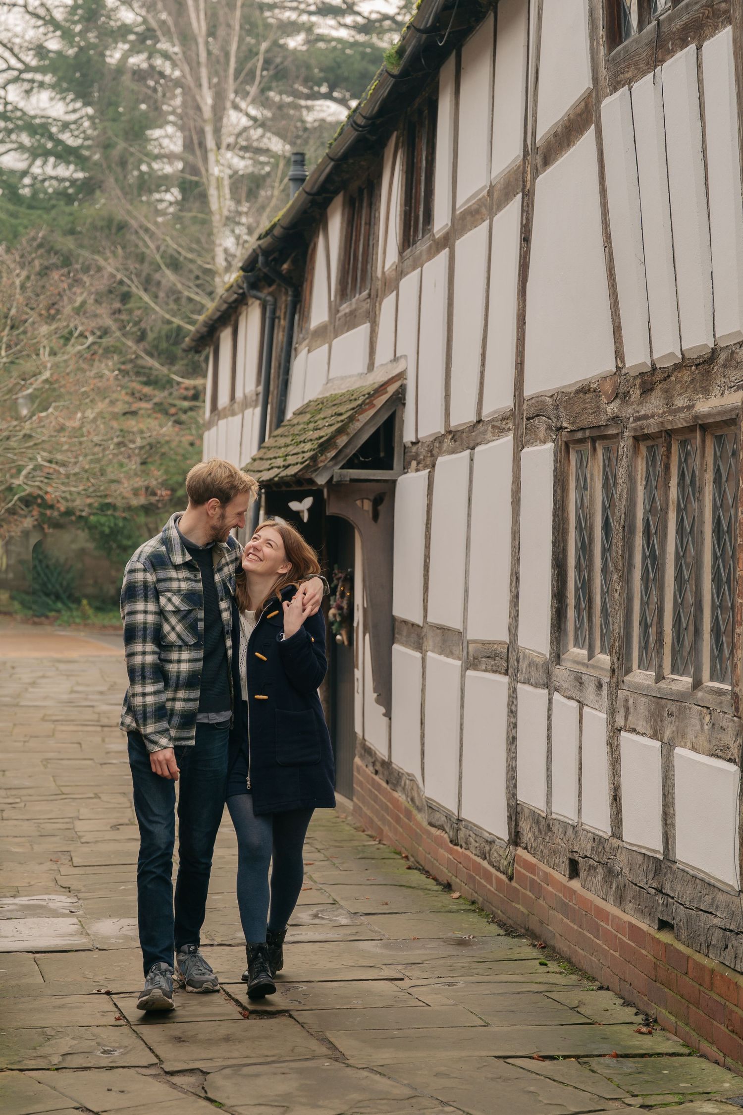 A couple walks together along a stone path beside a rustic half-timbered building in a romantic outdoor setting.