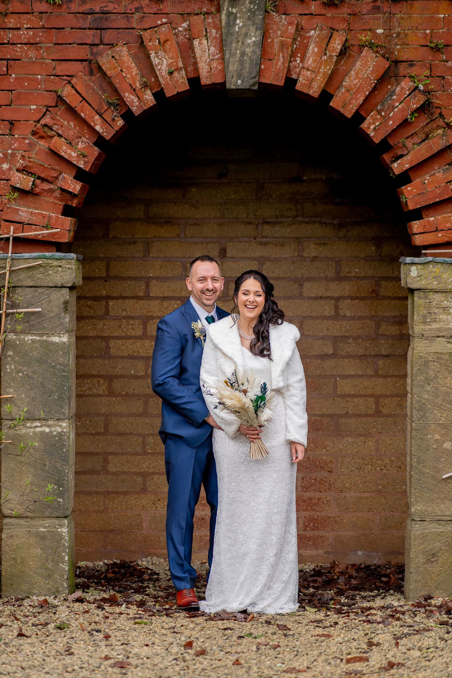 A wedding couple stands together under a rustic brick archway during an autumn day.