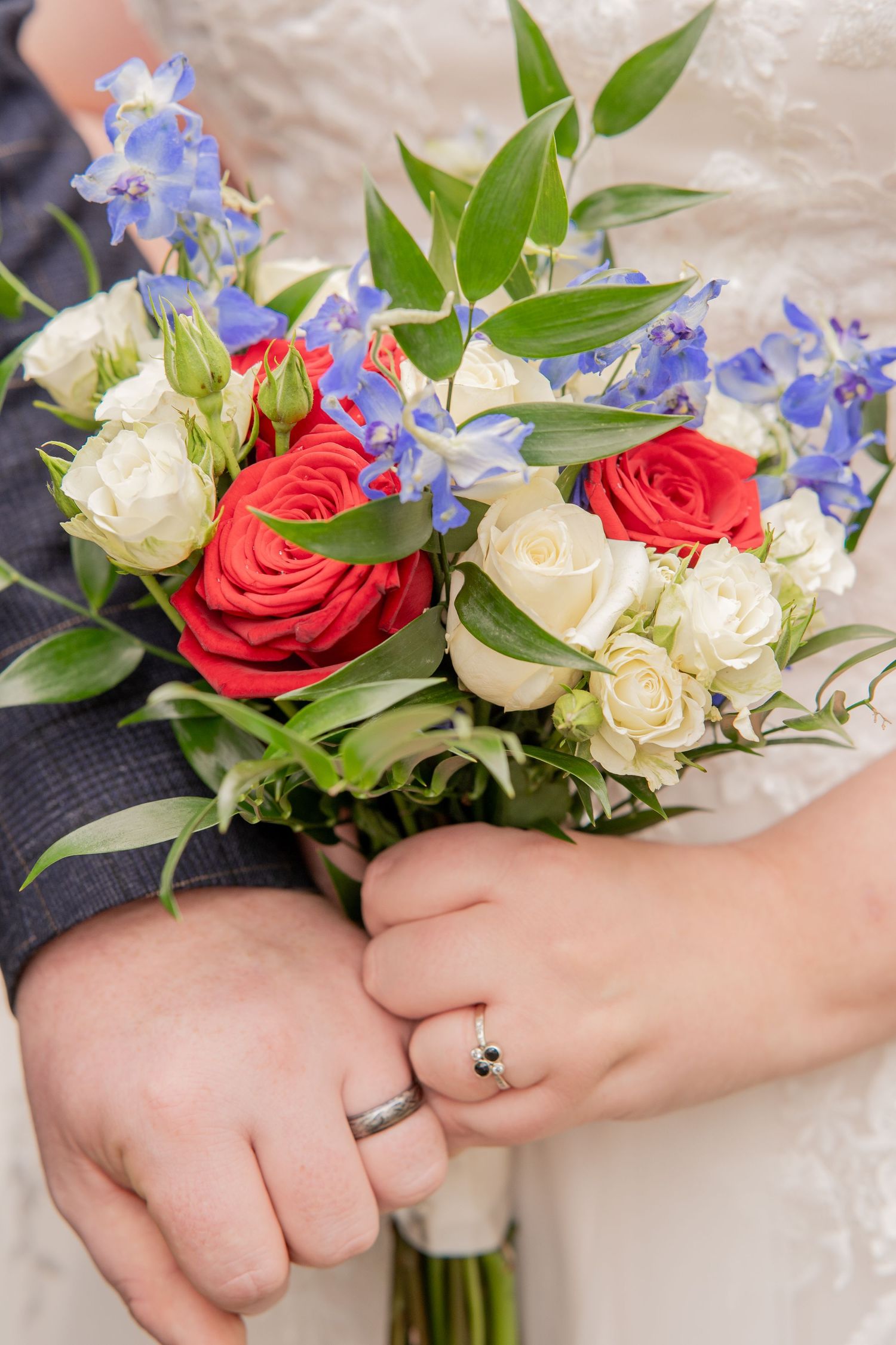 A wedding bouquet of red and white roses with delicate blue delphiniums while hands display an engagement ring.