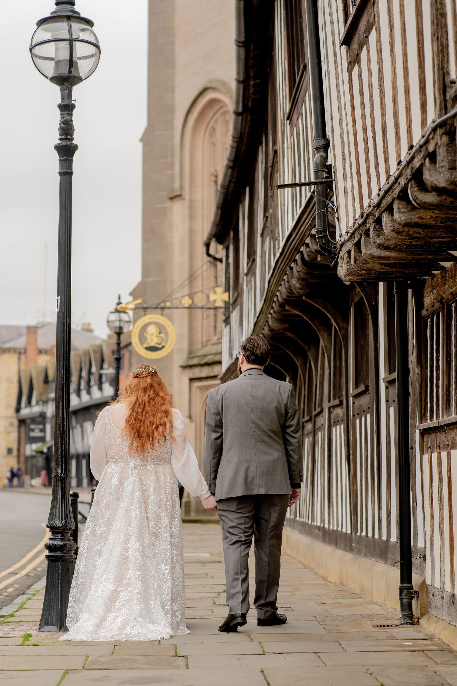 A couple walks down a historic Shakespearean  street with vintage lampposts and ornate architecture on their wedding day.