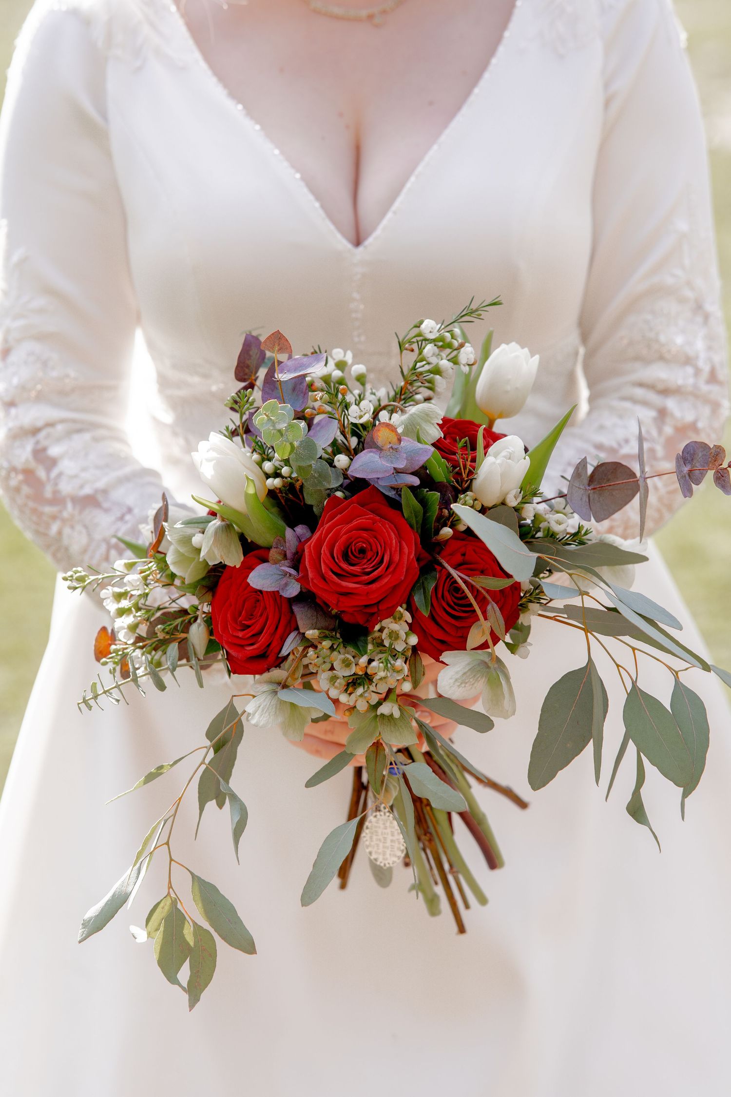 A wedding bouquet with red roses, white tulips, and eucalyptus leaves held against a white lace dress.