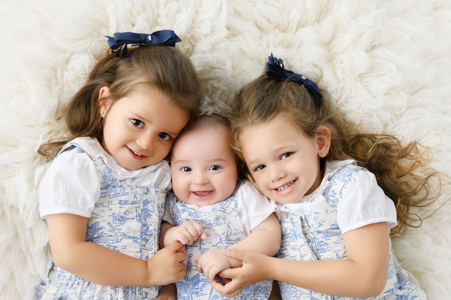 Three young siblings, two toddler girls with blue bows and a smiling baby, pose together on a fluffy white blanket.
