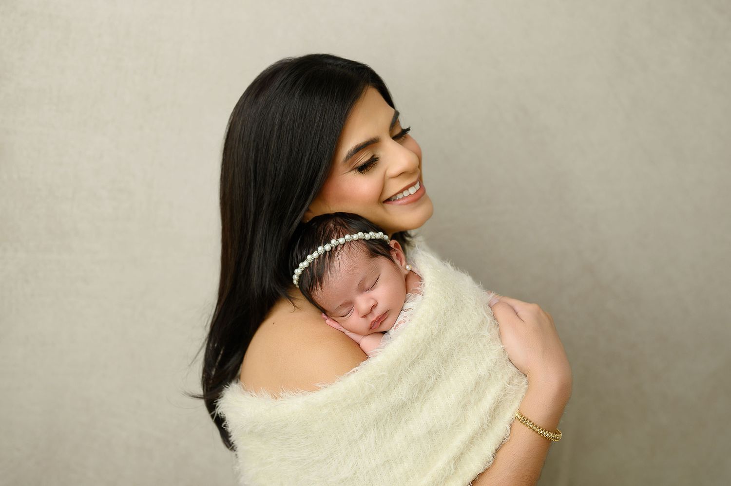 Smiling mother holding sleeping newborn baby wrapped in white, studio portrait photography.