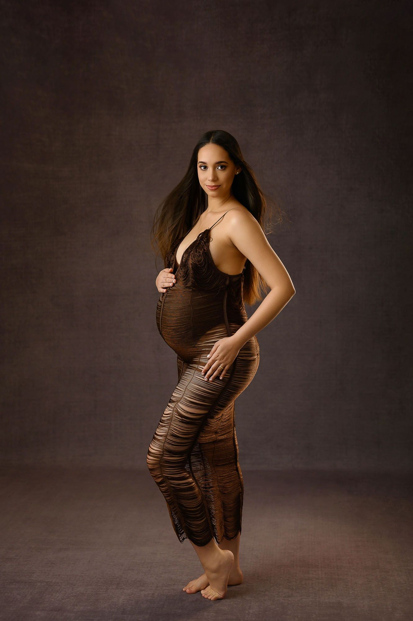 Pregnant woman in brown lace maternity gown posing elegantly against a dark studio backdrop.