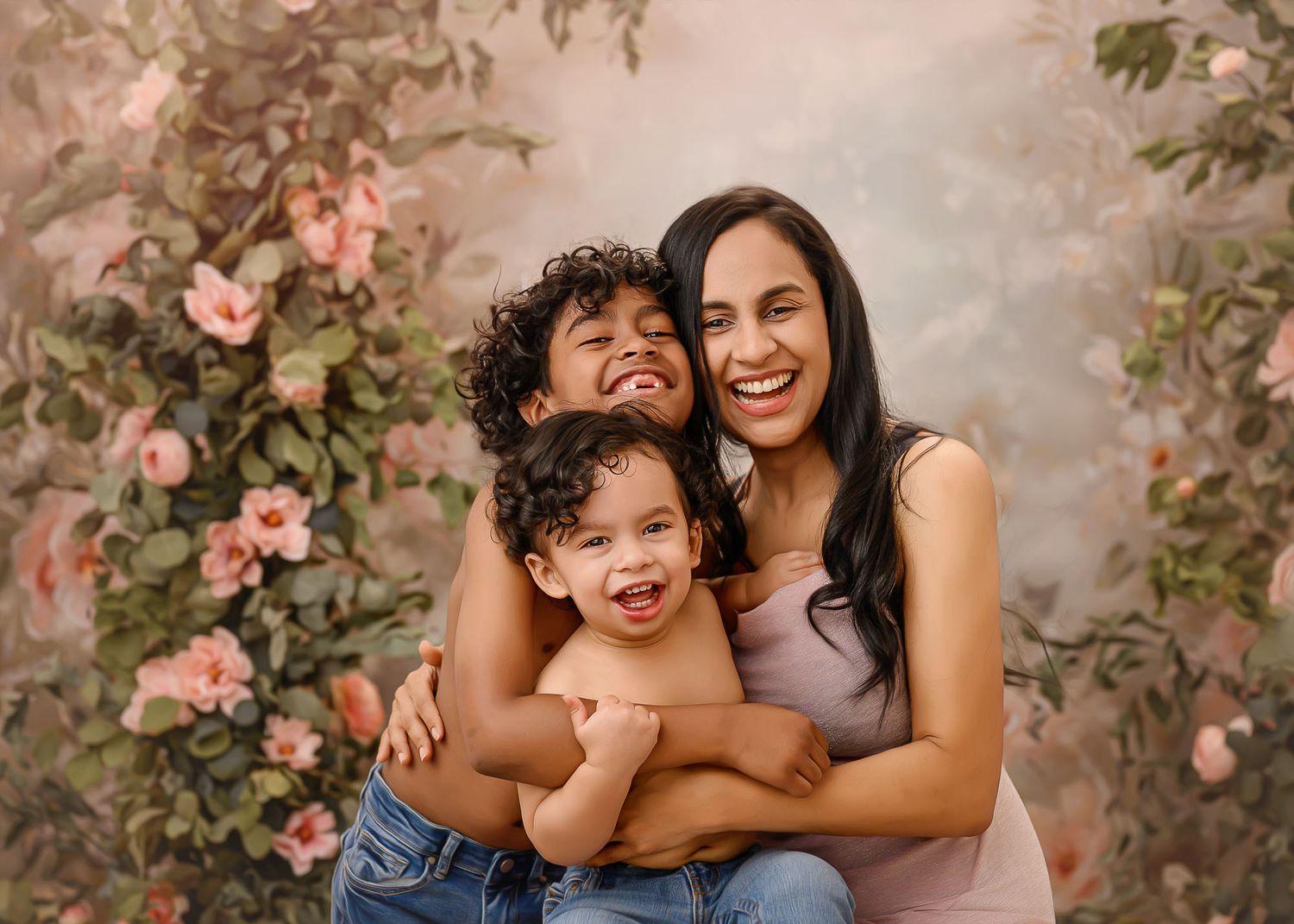 A joyful mother hugs her two young sons in front of a floral backdrop with pink roses.