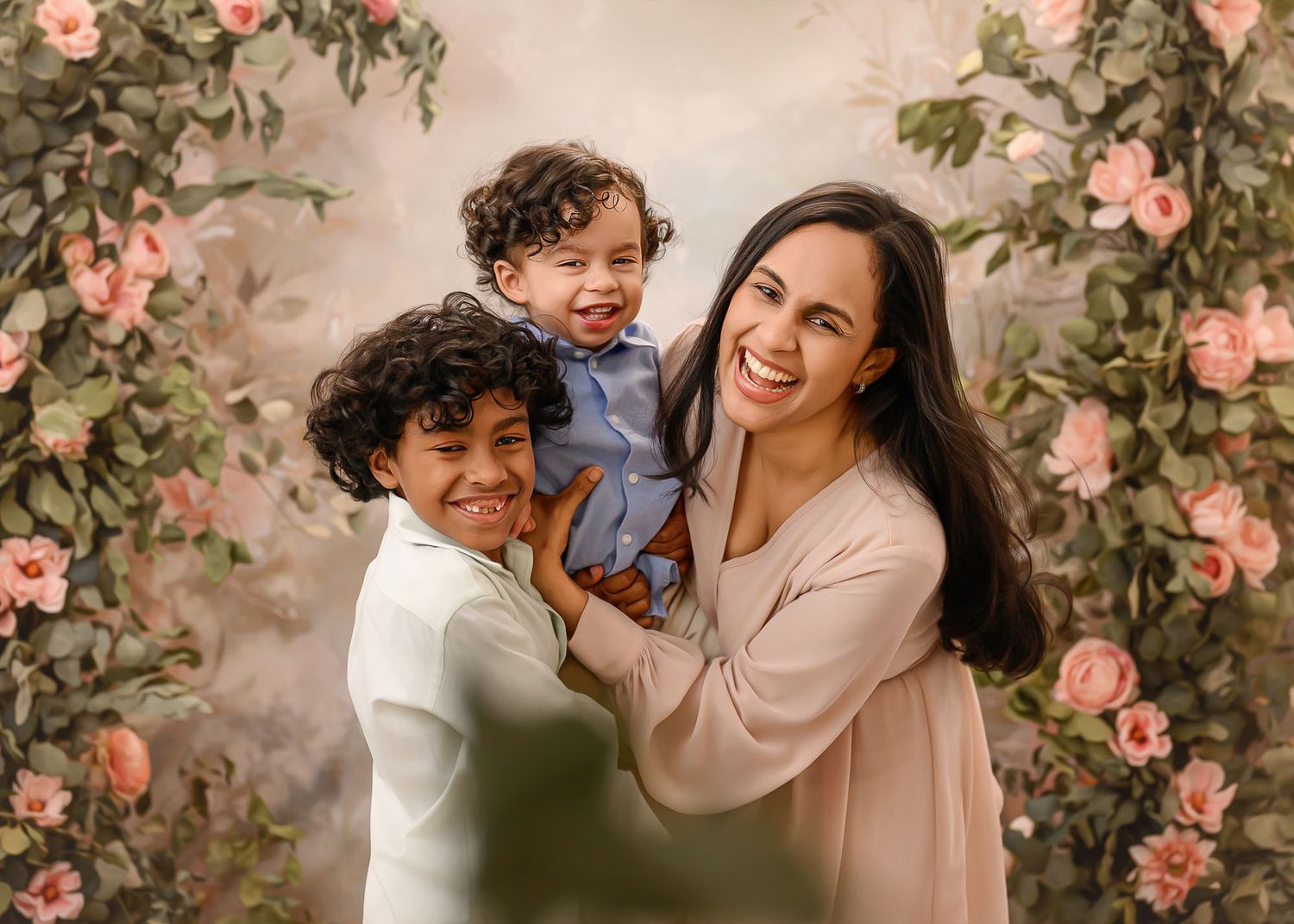 A smiling mother holds two young children in front of a floral backdrop with pink roses.