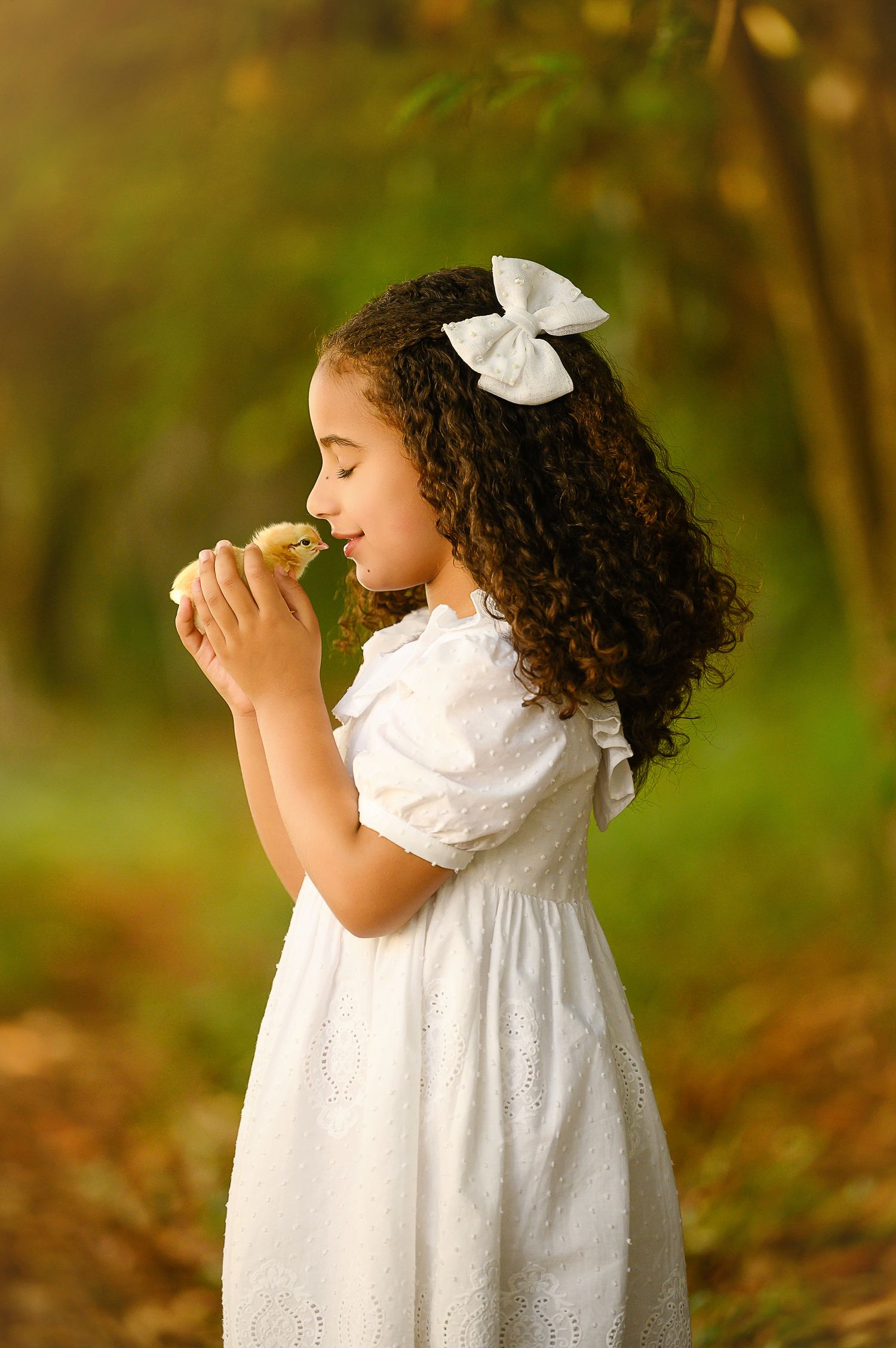 A young girl in a white dress and bow holds a baby duckling to her face in a sunlit forest setting.