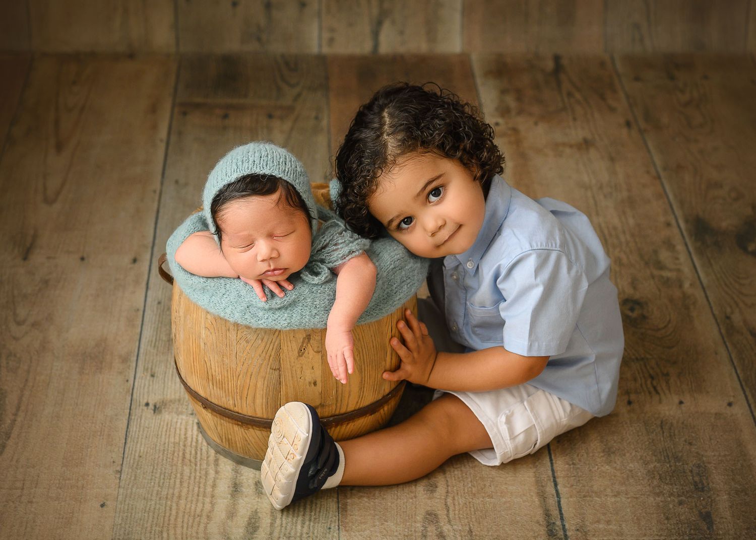 Toddler boy sits beside a wooden barrel holding a sleeping newborn baby wrapped in teal knit on a wood floor.