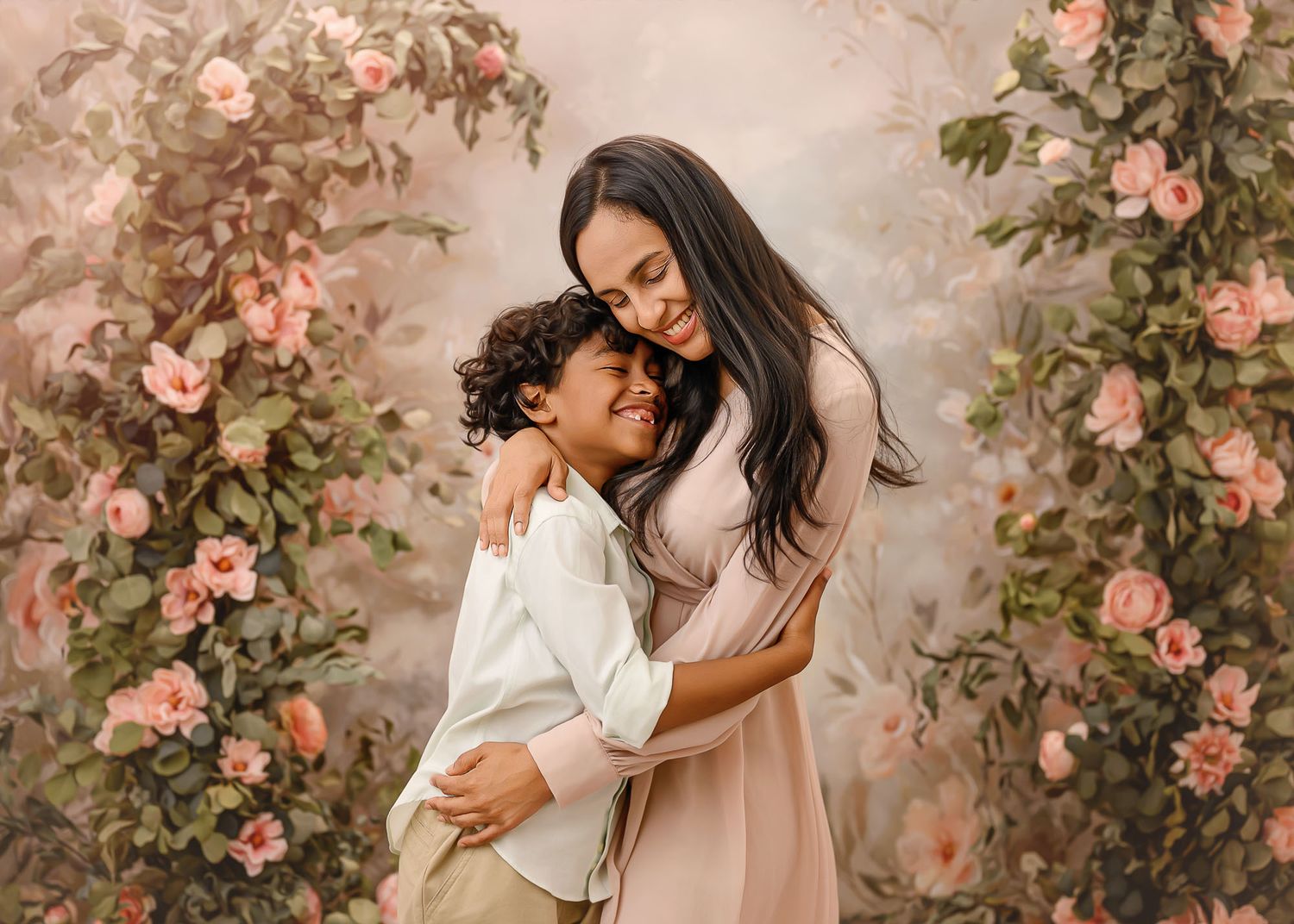 A mother in a blush dress tenderly embraces her young son before a lush floral arch backdrop.