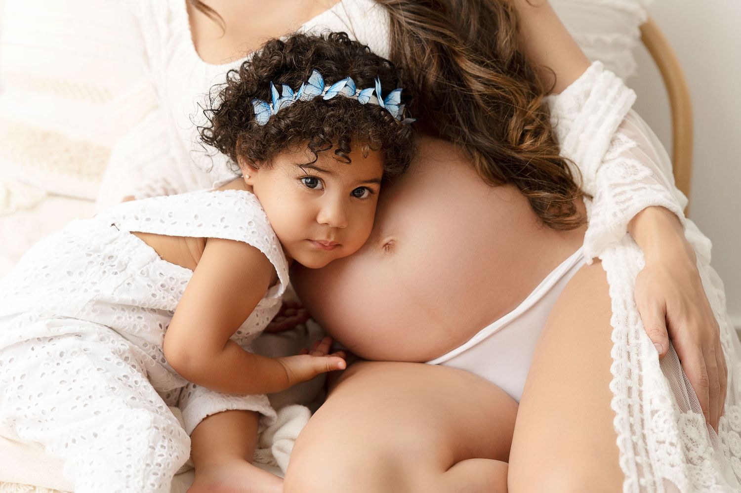 A toddler girl with curly hair rests her head on her pregnant mother's bare baby bump during a maternity photoshoot.