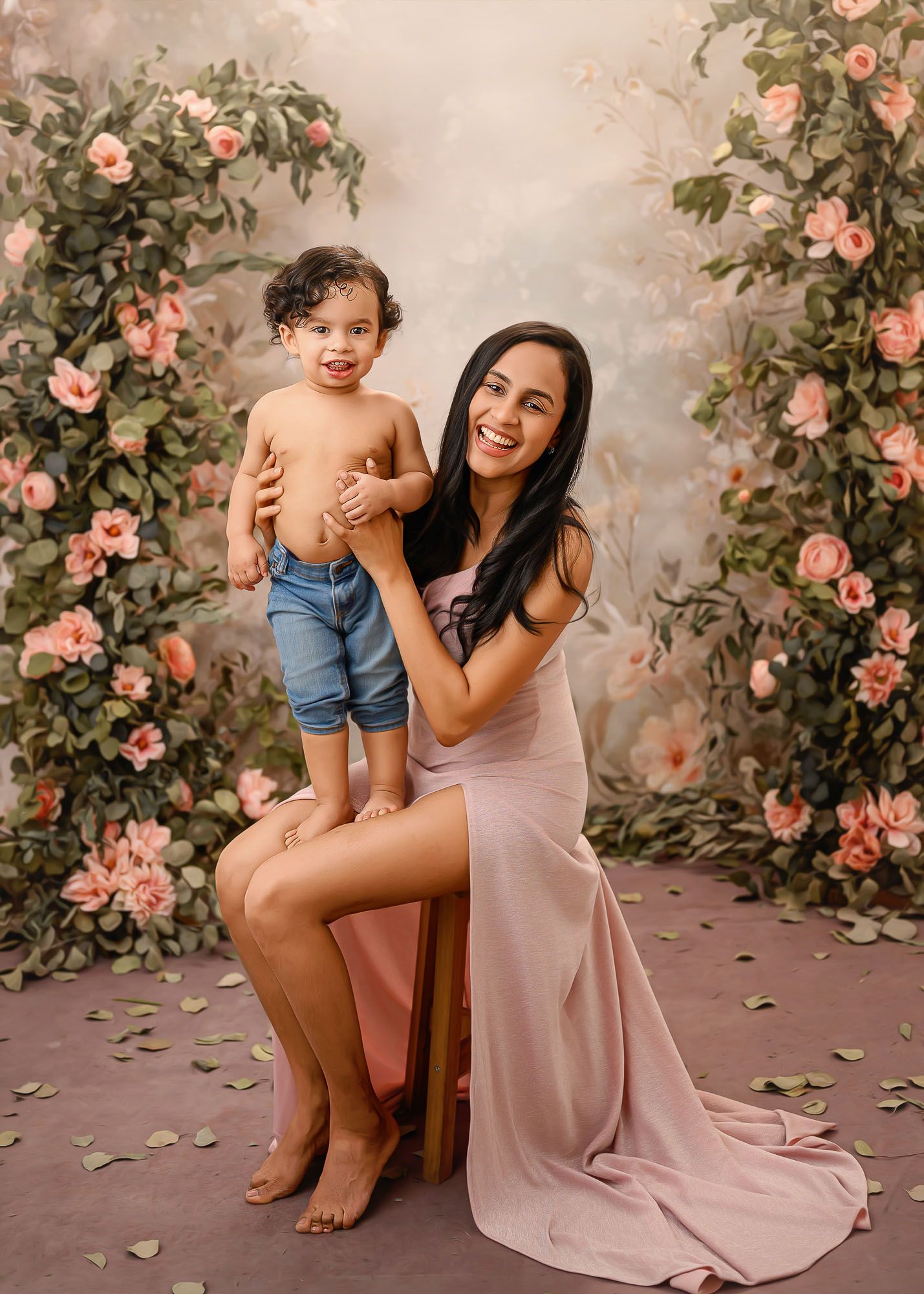 Woman in pink gown holds toddler boy in denim shorts before floral rose arch backdrop.