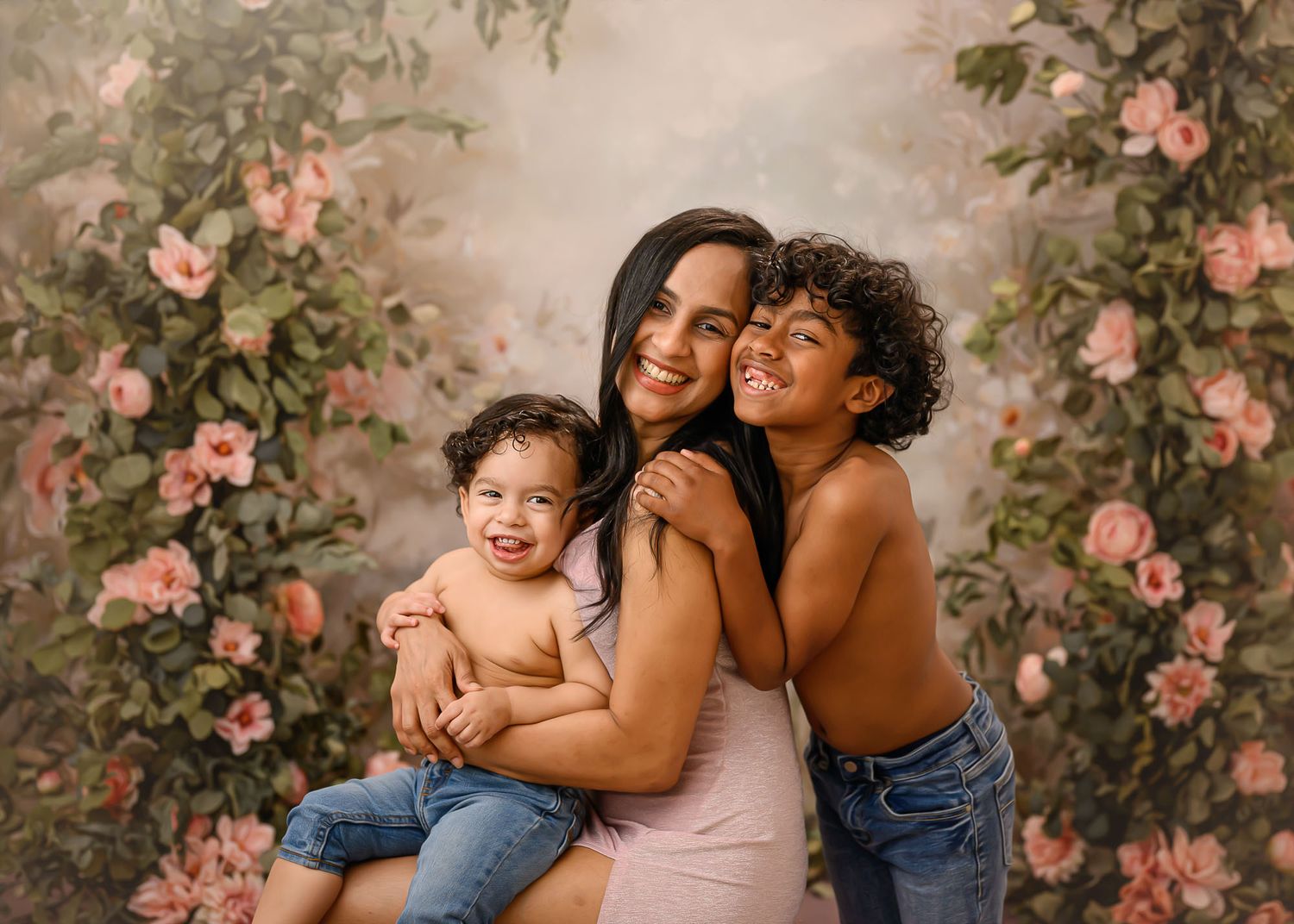 A mother smiles warmly holding her baby and young son against a floral backdrop in a professional family portrait.