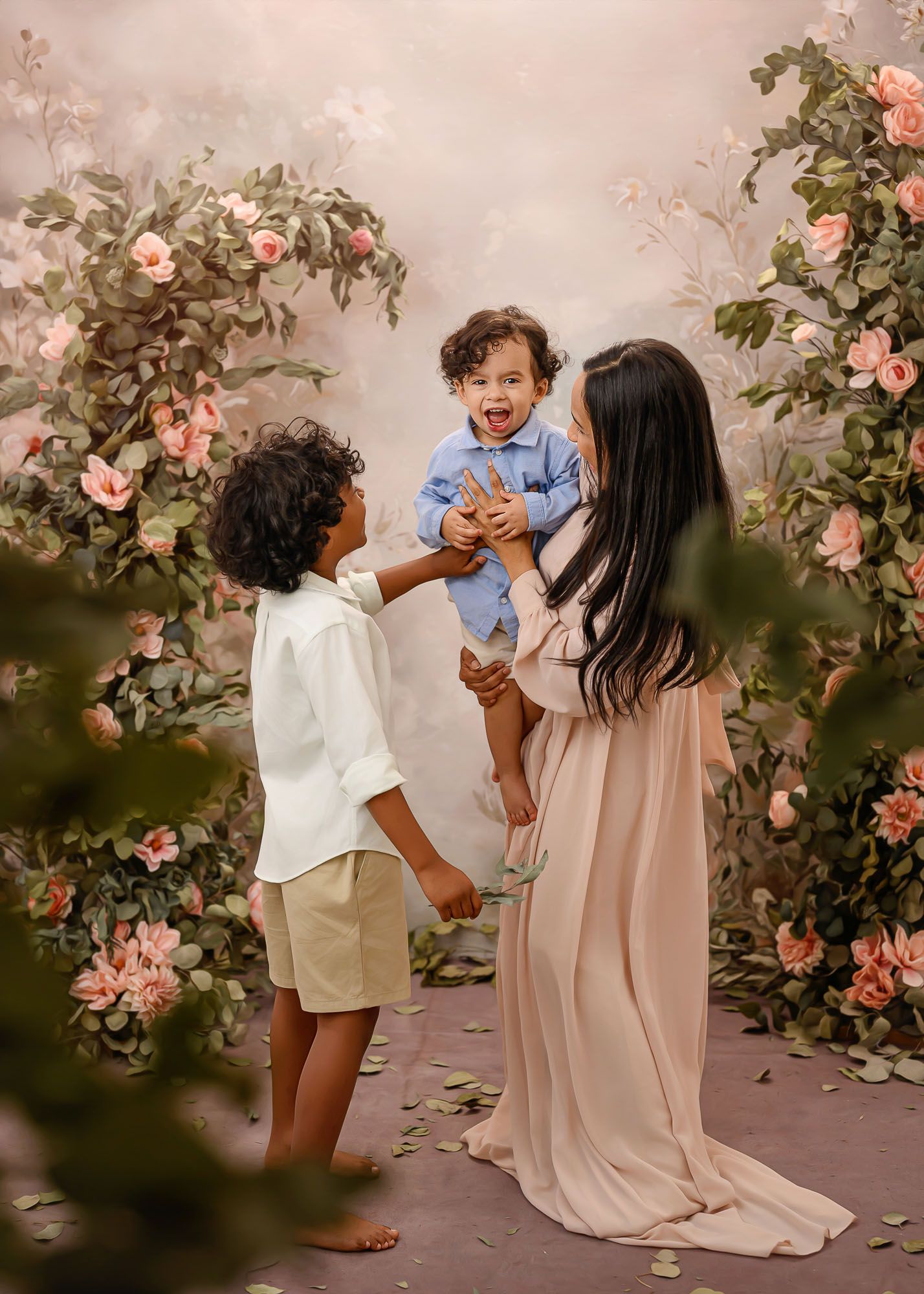 A mother holds her laughing toddler while her older son reaches up amid a floral arch backdrop.