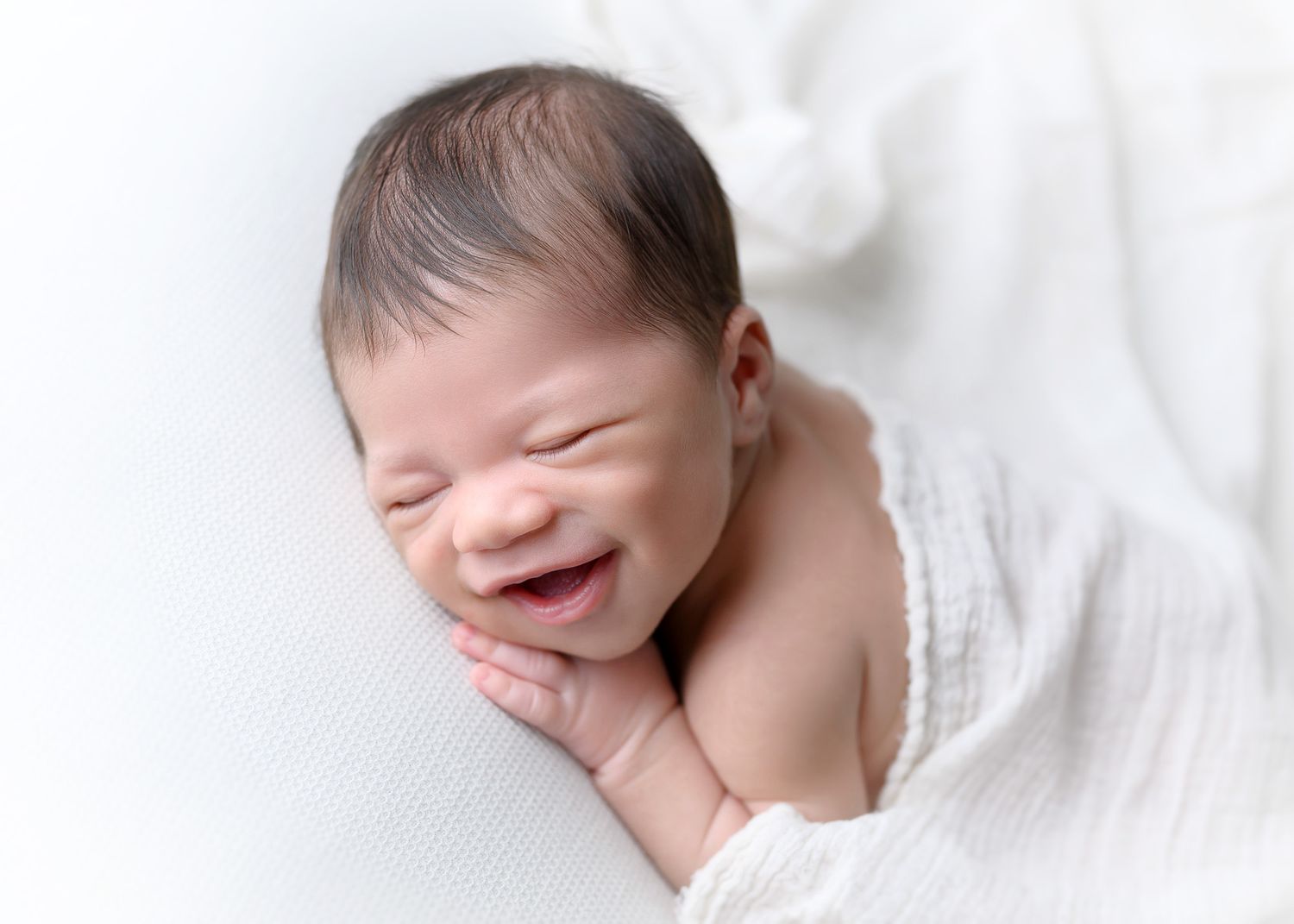 Smiling baby lying on white blanket with hand near face.