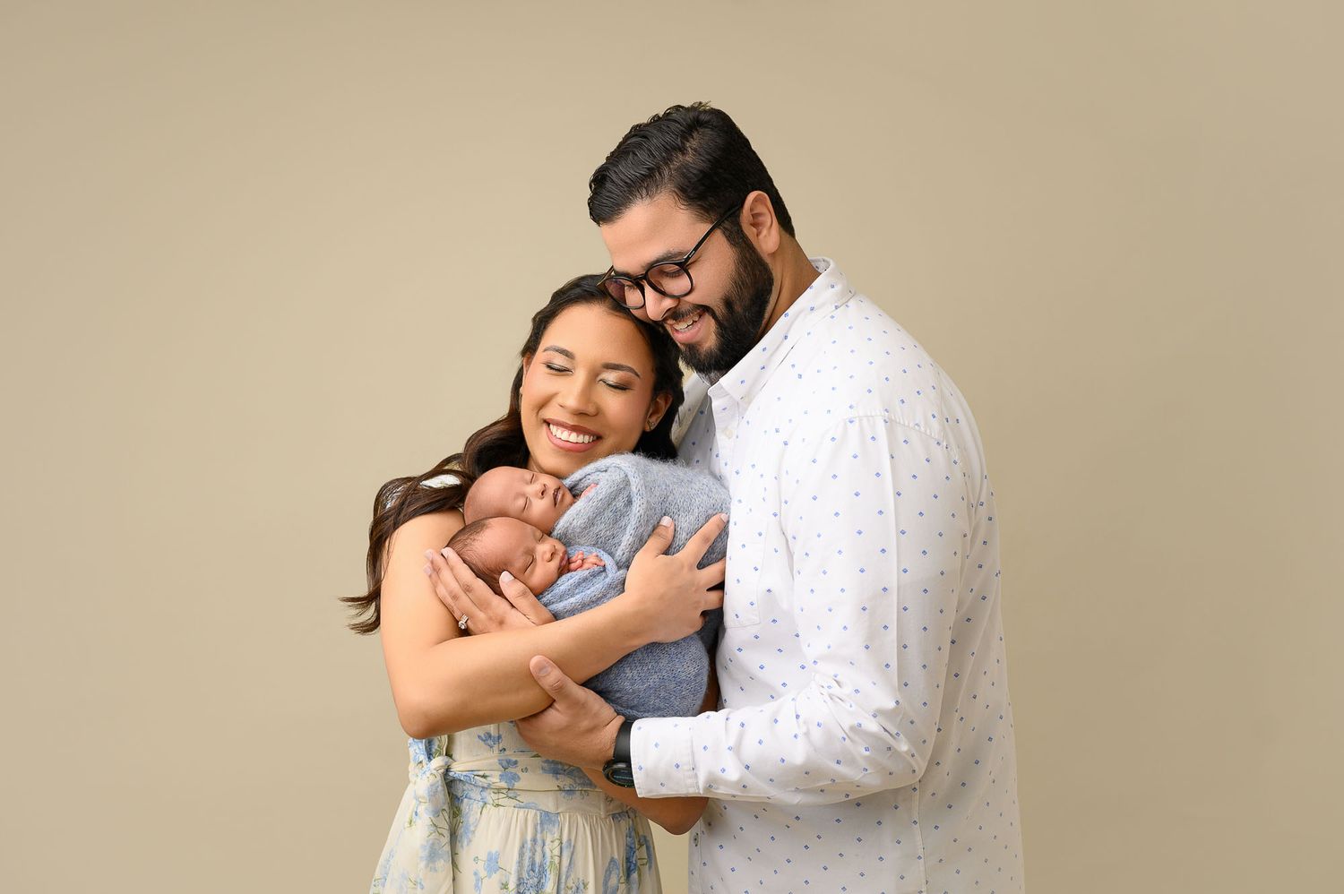 A couple tenderly holds their sleeping newborn baby, posing for a professional portrait against a neutral beige background.
