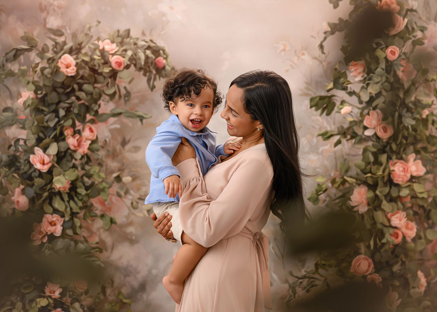 A mother in a blush dress holds her smiling baby boy before a floral arch with pink roses.
