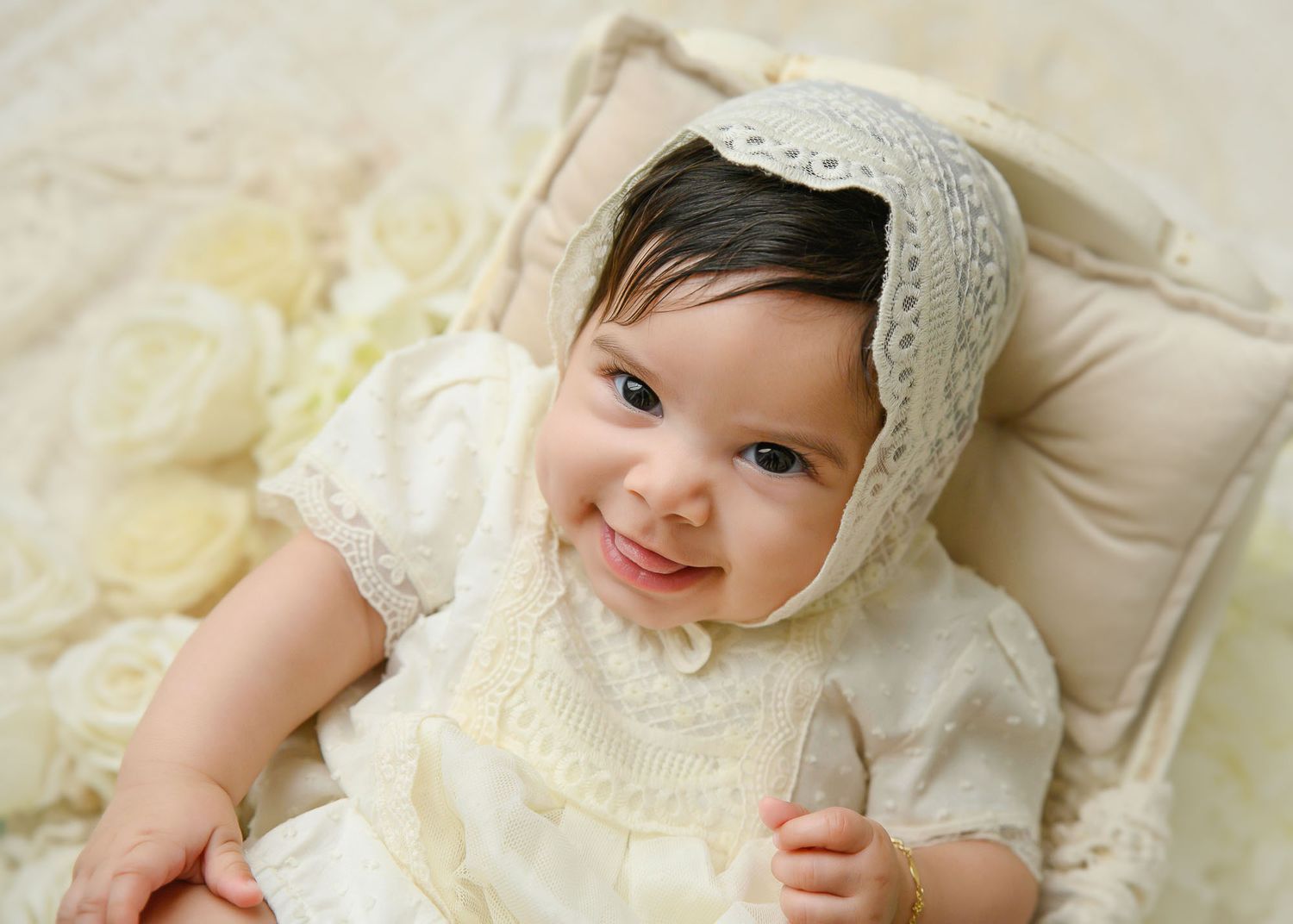 Smiling baby in cream lace bonnet and dress lying on soft ivory bedding with roses.